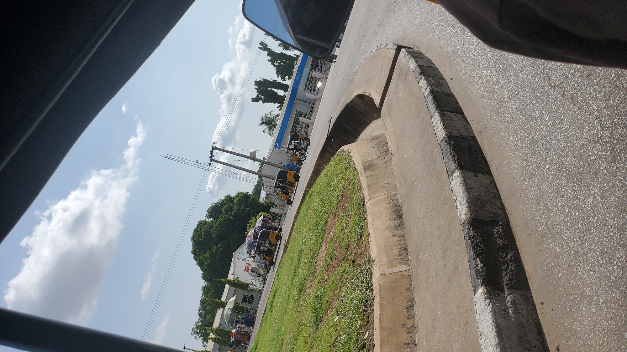 The image, taken from a moving vehicle, shows a street scene in Jalingo, Nigeria. The foreground features a concrete road with a distinctive curb, painted with alternating black and white stripes, indicating a traffic calming measure or a boundary. To the left, a grassy median curves away from the road, bordered by the same striped curb.
In the background, several yellow auto-rickshaws, known locally as "Keke Napep" or "Keke Marwa," are visible. Some are parked, while others appear to be in motion. One auto-rickshaw is laden with umbrellas, suggesting it may be a mobile vending unit or transporting goods. Another is occupied by passengers. A person on a motorcycle is also present.
Buildings line the street, including what appears to be a bank with the UBA logo prominently displayed. Green trees and power lines are scattered throughout the scene, contributing to the urban landscape. The sky is partly cloudy with a bright, sunny atmosphere, indicating daytime. The overall impression is a typical street scene in a Nigerian city, with a focus on local transportation and commerce.