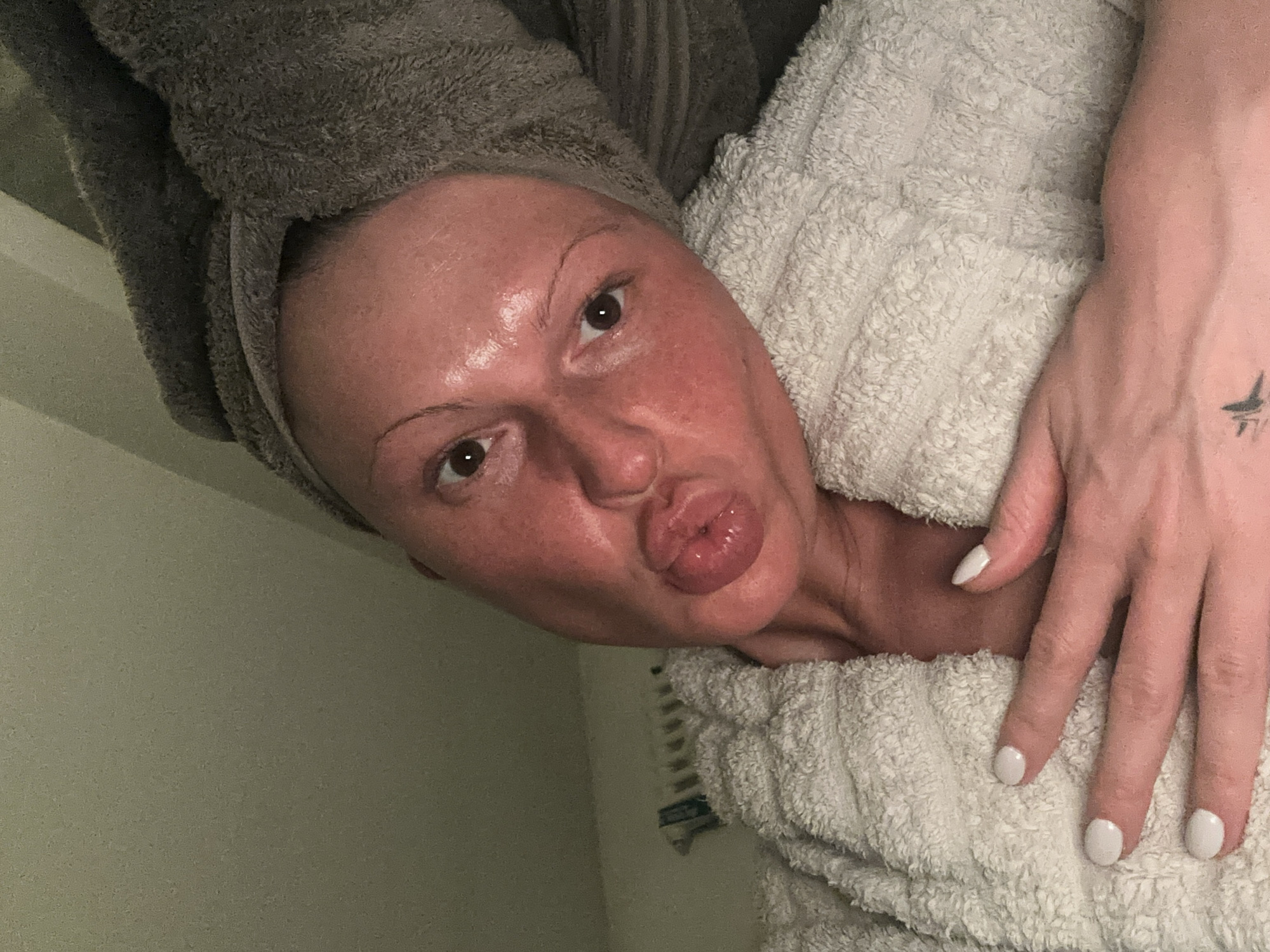 An adult female is depicted in a close-up, upward-angled selfie, within an interior location, presumably a residence in Tunbridge Wells, United Kingdom. She wears a grey terrycloth towel wrapped around her head and a white terrycloth bathrobe. Her face appears flushed red and exhibits a moist, reflective sheen. Her eyes, with dark irises, are open and directed at the camera. Her eyebrows are light. Her mouth is pursed into a pronounced pout. Her left hand rests on her chest, displaying fingernails painted white. A small, dark, stylized bird-like tattoo is visible on the back of her left hand near the wrist. A white, finned radiator is partially visible in the background against a light-colored wall.