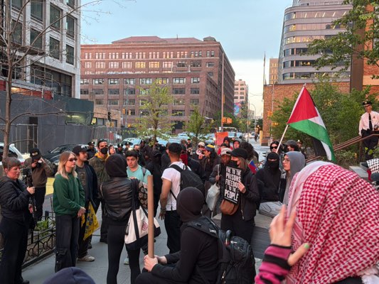 A daytime outdoor scene shows a group of approximately 30 people gathered on a city street. The majority of people appear to be in their teens to middle age. Most of the people are dressed in casual clothing, and some have face coverings. One person is holding a sign, a message which cannot be determined. In the background, a Palestinian flag is being held aloft. A man in a police uniform is visible on a set of steps to the right of the crowd. The setting appears to be an urban area with buildings in the background. The image has a natural daylight quality, suggesting it was taken in the late afternoon. The people are interacting with each other and appear to be engaged in a demonstration.
The scene is likely in Hoboken, New Jersey, United States.