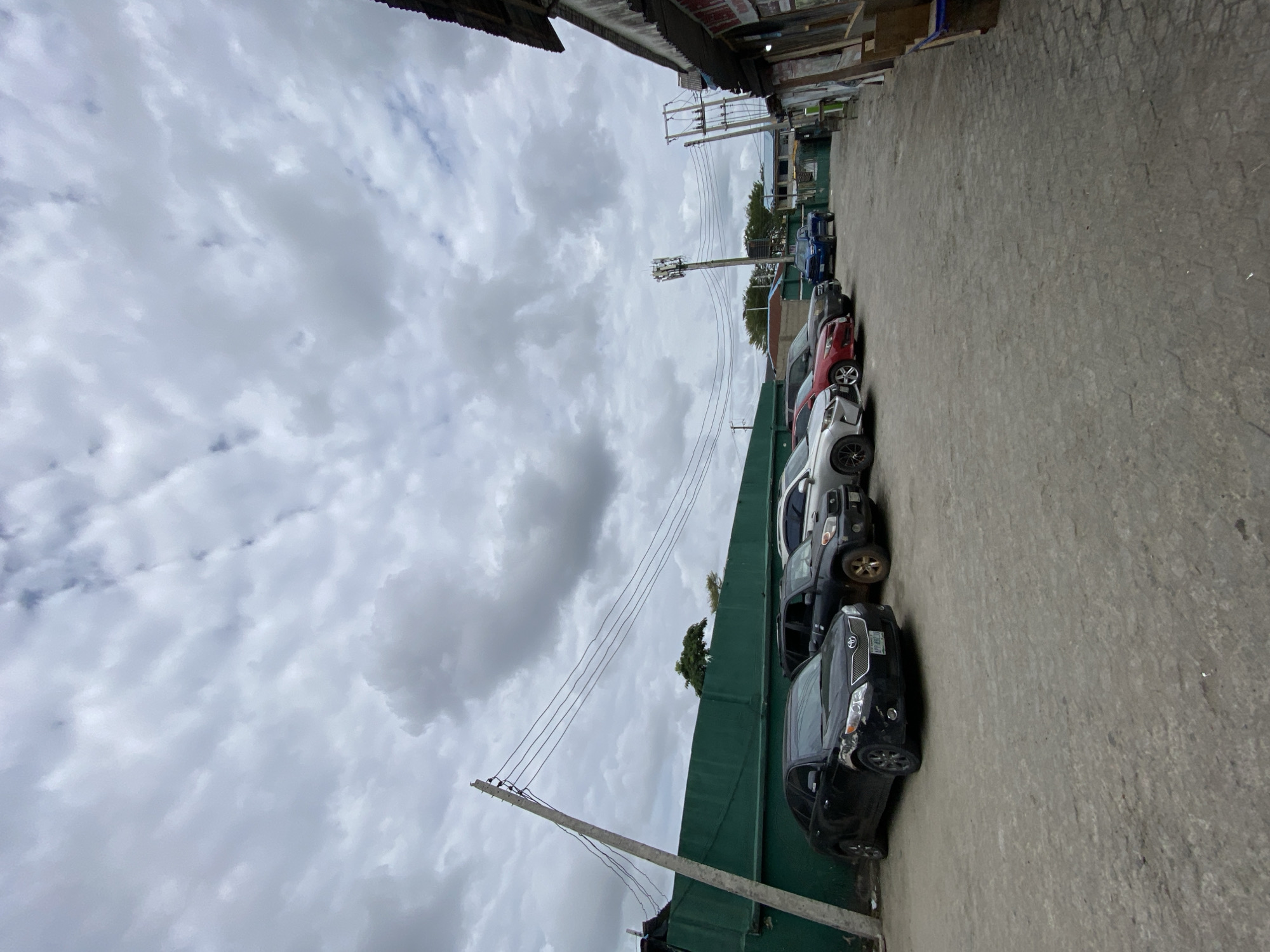 The scene is an urban street in Ado, Nigeria, lined with parked cars along a green wall. A utility pole with power lines stretches across the sky, which is overcast with heavy clouds. Buildings are visible on either side of the street, constructed from corrugated metal and wood. The ground is paved. There are no people visible.