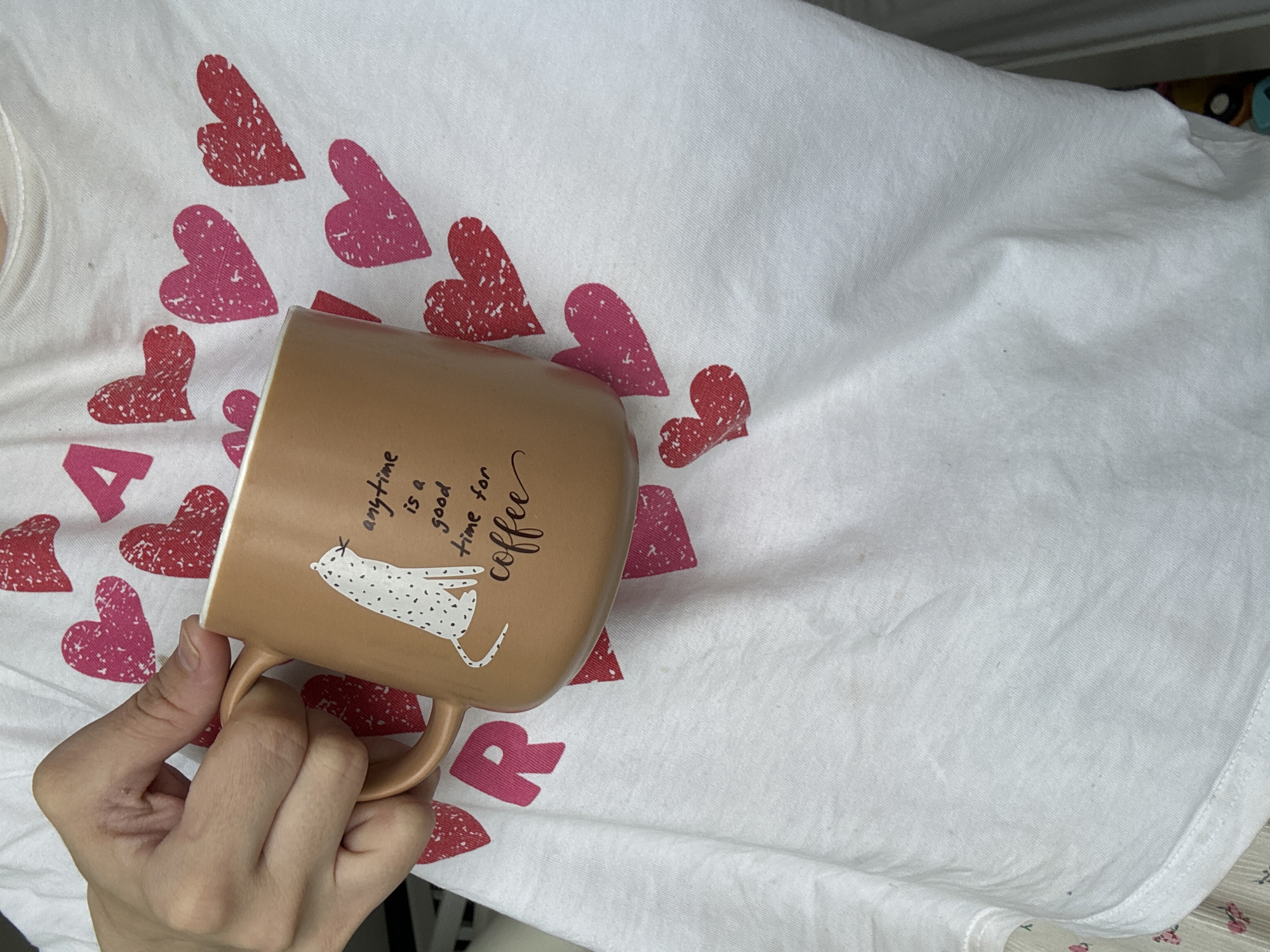 The image presents a close-up, top-down view of a hand holding a light brown, matte mug above a white fabric surface. The fabric is adorned with numerous red and pink heart patterns, some appearing speckled. Partially visible letters on the fabric include a large "A" and "R", suggesting a word like "HEART" or "LOVE" in combination with the hearts.
The mug features a white, spotted cat lying down on its side, with the phrase "anytime is a good time for coffee" written above it in a decorative script. The person's hand is visible in the lower-left, grasping the mug's handle. The scene takes place indoors. This activity is occurring in Chaikovsky, Russia.