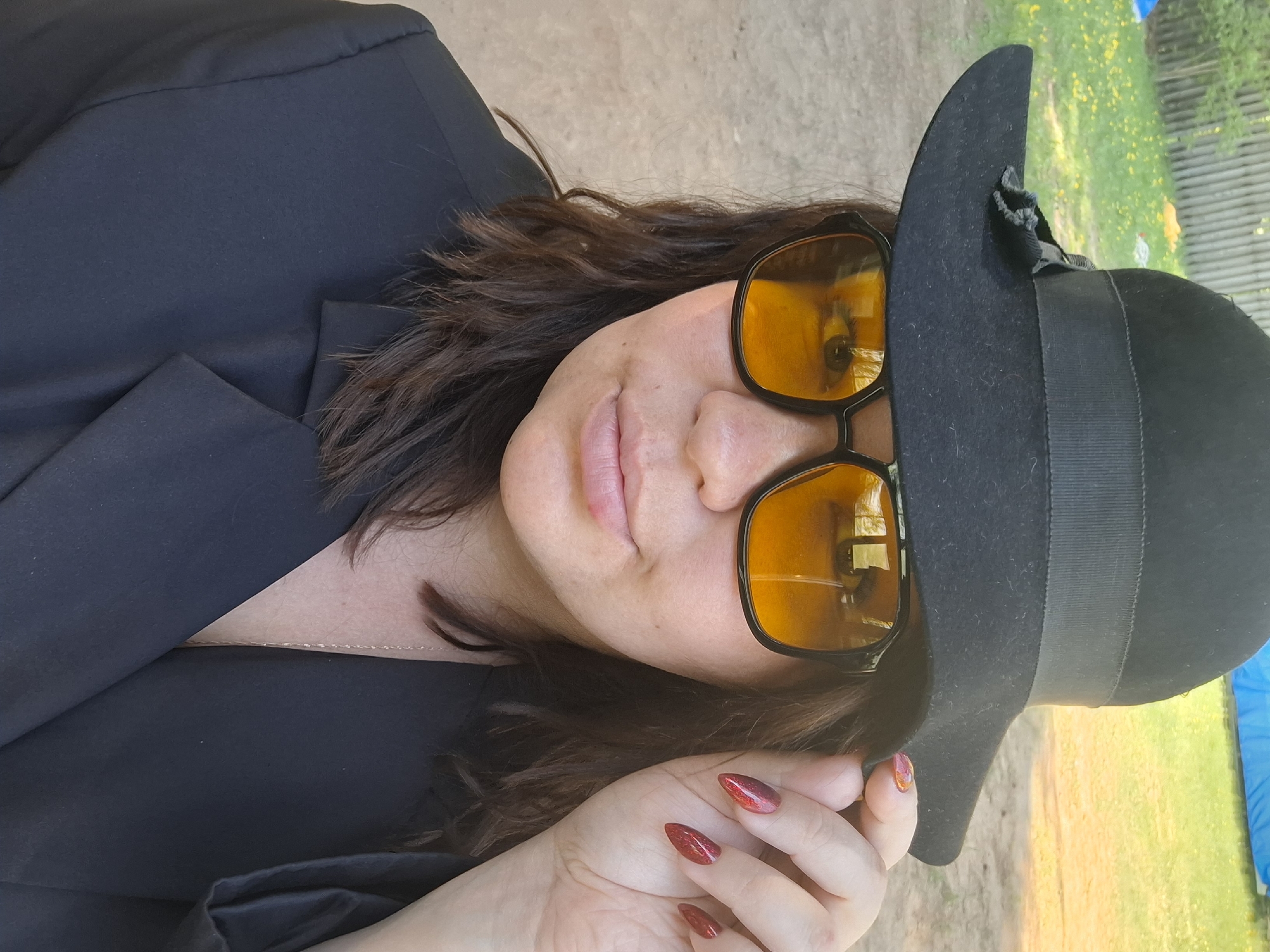 A woman is captured in a medium shot, wearing a black blazer, a black hat adorned with a ribbon, and yellow-tinted sunglasses. The hat partially obscures her face. Her fingernails are painted with a red, glittery polish.
The background appears to be an outdoor setting, possibly a park or garden. Vegetation, including grass and foliage, is visible in the background, suggesting a natural or semi-natural environment. The presence of a yellow and green colored picnic table in the back suggests the location is a public park, or possibly in a garden in Chaikovsky, Russia. The image is rotated.