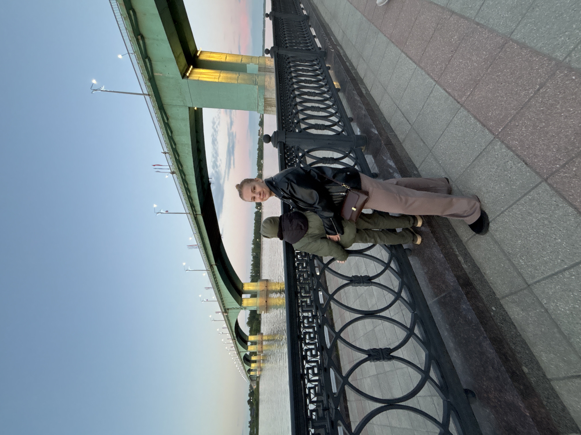 A family is photographed on the embankment along the left bank of the Oka River in Rusanovo, Russia, in front of the Kanavinsky Bridge. A woman wearing a black leather jacket and beige pants stands next to a young child who is wearing a green hooded jacket. The woman looks at the camera. They are standing next to a black decorative iron fence. In the background, the Kanavinsky Bridge crosses the Oka River under a clear, early evening sky. Lampposts line the bridge. The bridge is visible in the water.