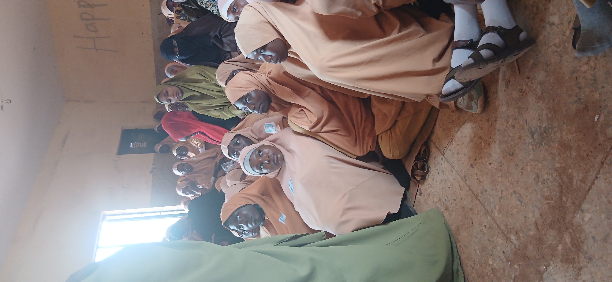 A group of young women and girls wearing hijabs and traditional Islamic attire are seated on the floor of a room that appears to be a classroom or community hall. The lighting suggests it is daytime, with light streaming in from a window on the left side of the frame. The walls are painted a light color and show signs of wear, with some graffiti or markings visible, including the word "Happy" written in large letters on the upper right wall. The floor is tiled with a speckled pattern. Some individuals are looking towards the left, possibly at someone addressing the group, while others are looking forward or down. One person in the foreground is wearing a light peach-colored hijab and is looking upwards. Another person next to them is also in a peach-colored hijab and looking forward. Towards the back, several individuals are visible, including one in a red hijab and another in a dark brown or black hijab and niqab. Some of the individuals are wearing sandals. The scene conveys a sense of gathering or instruction. The location is identified as Hayin Banki, Nigeria.