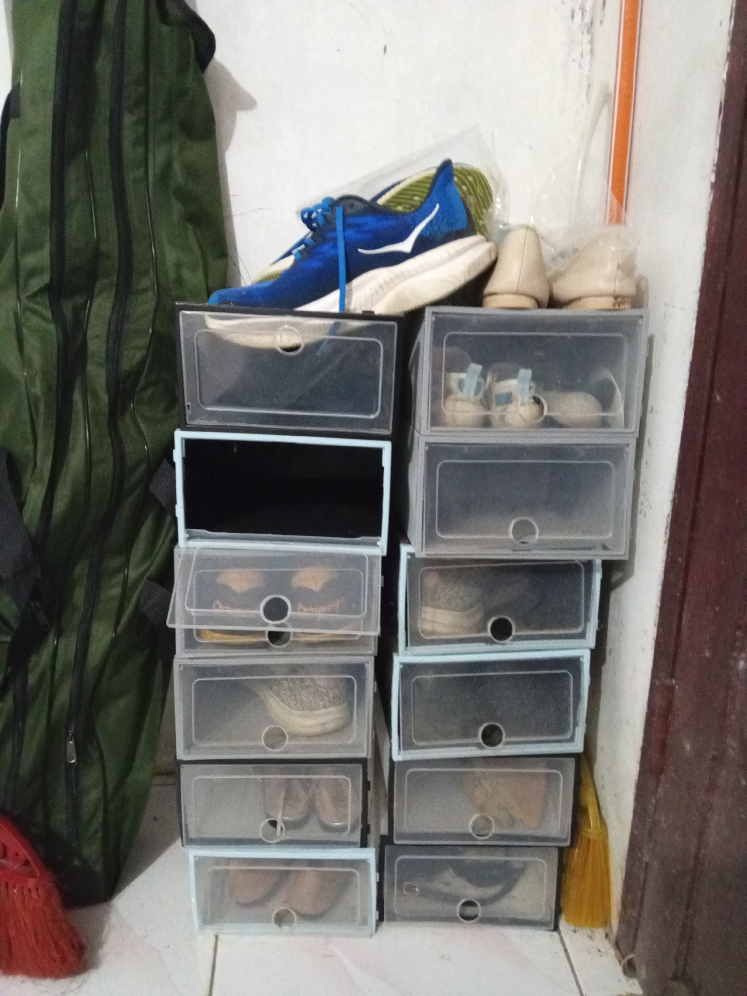 In Palohigeuh, Indonesia, a stack of clear plastic shoe boxes is visible. The boxes are arranged vertically, with some containing shoes. The top box has a blue and white athletic shoe. Below that, two small light-colored shoes are present. Further down, other shoe boxes contain pairs of shoes in various colors and styles. A large green bag stands to the left of the shoe boxes, and a yellow broom leans against the wall on the right. The scene appears to be indoors, with a tiled floor and a white wall.