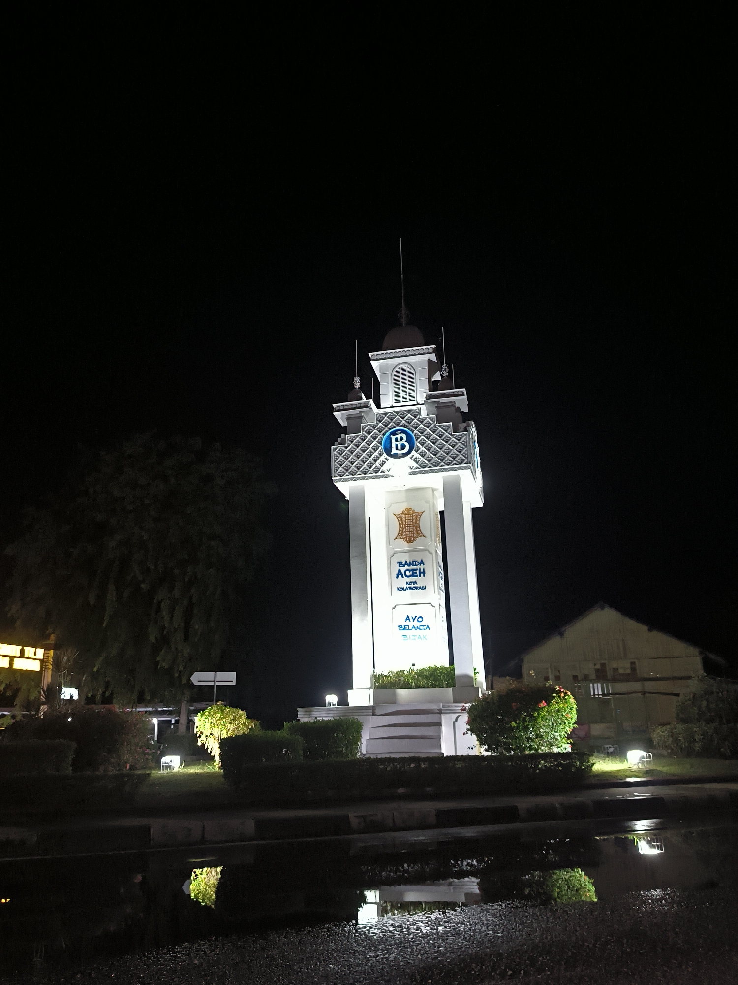 This nocturnal photograph captures a brightly illuminated, white tower-like monument in Banda Aceh, Indonesia. The central structure, resembling a stylized clock tower or landmark, features intricate patterns near its top and a small dome. A blue "B" logo within a circle is prominent on one side near the apex, and below it, an emblem in gold and white is visible. Further down, the tower displays the text "BANDA ACEH" and "KOTA KOLABORASI" (Collaboration City), with "AYO BELANJA BIJAK" (Let's Shop Wisely) written near its base.
The tower is surrounded by well-maintained low hedges and bushes, some of which are lit by ground-level spotlights, casting a soft glow. To the left of the monument, a large, dark tree stands, mostly silhouetted against the black night sky, with some faint illumination emanating from below its canopy. On the right, a simple, light-colored building with a peaked roof is partially visible and dimly lit.
The foreground consists of a dark, wet surface, likely pavement or a road, which prominently displays reflections of the tower's bright lights, the illuminated bushes, and other unseen light sources. These reflections suggest that it has recently rained, contributing to the glossy appearance of the ground. The sky is completely dark, confirming it is nighttime. There are no people visible, and the scene conveys a serene, quiet atmosphere in an urban setting.