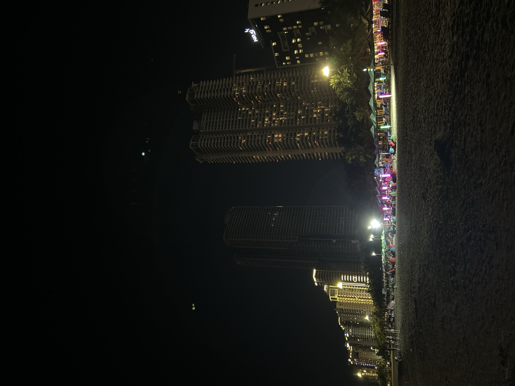 At night on a sandy beach in Da Nang, Vietnam, numerous people are seen relaxing on beach chairs under colorful umbrellas. The scene is illuminated by the lights of several high-rise buildings that line the coast, their windows glowing. A prominent building displays the "WYNDHAM" logo. The sand is dark and shows footprints. The sky above is black with a few distant lights visible.