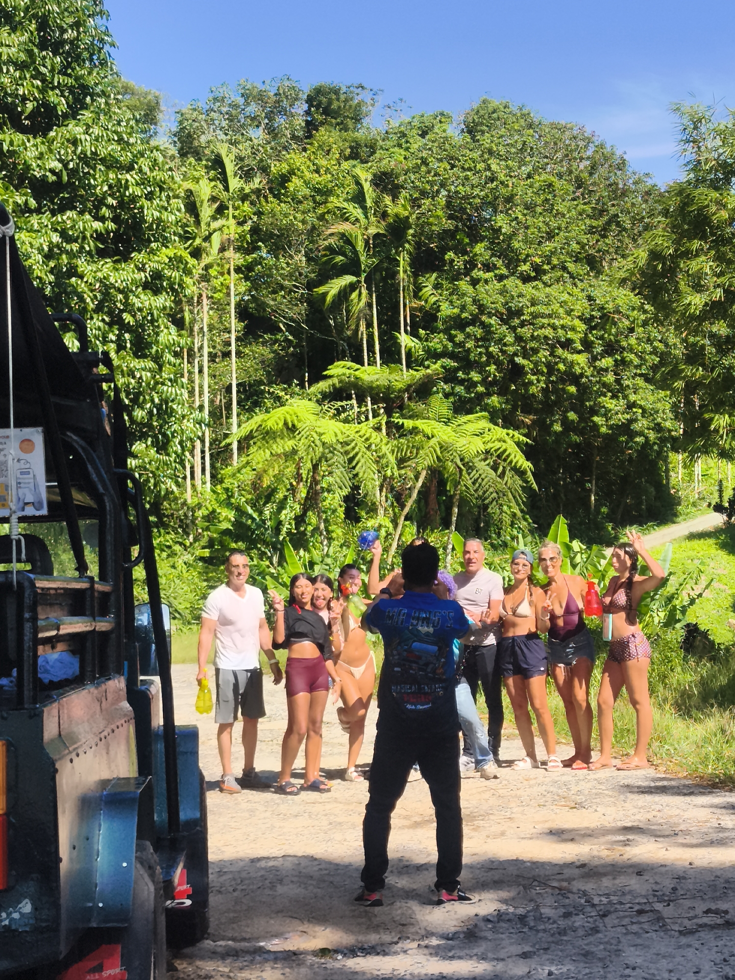 An outdoor group photograph is taken in a jungle setting on a sunny day. A group of nine individuals, eight in the foreground and one positioned slightly behind them, are posing for a photo. The group consists of a mix of men and women, all dressed in casual attire, including swimsuits, shorts, and t-shirts. The individuals appear to be in good spirits, with some smiling, making peace signs, and holding water guns.
To the left of the frame, a portion of an open-top safari-style vehicle is visible. The vehicle is dark-colored and appears to be a mode of transport. The background is dominated by lush green vegetation, including tall palm trees and dense foliage, indicative of a tropical environment. A dirt road or pathway is visible behind the group. The sky above is a clear, bright blue. The scene suggests a recreational activity or excursion in a natural, possibly humid, climate. Based on the flora and common tourist activities in the region, this scene could be located in the lush jungle areas of Ko Samui District, Thailand, possibly near a waterfall or adventure park.
