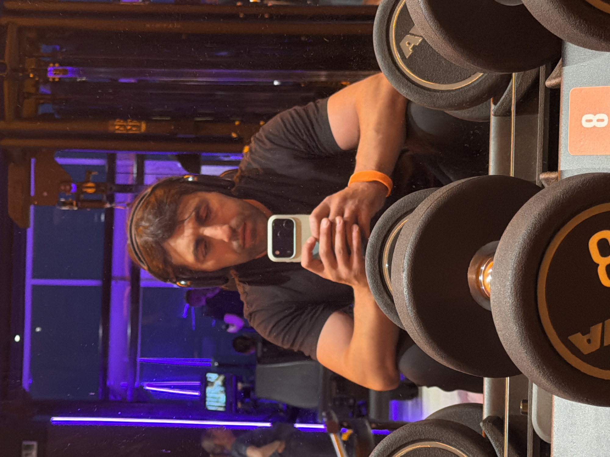The image shows a man taking a selfie in a gym. He is wearing headphones and an orange wristband. He is holding a white smartphone. A rack of dumbbells is visible in the foreground, with "8" printed on a label attached to the rack. The gym appears to have blue and purple neon lighting and mirrored walls. There is no visual evidence to suggest that the location is in the city of Krasnoyarsk, Russia.