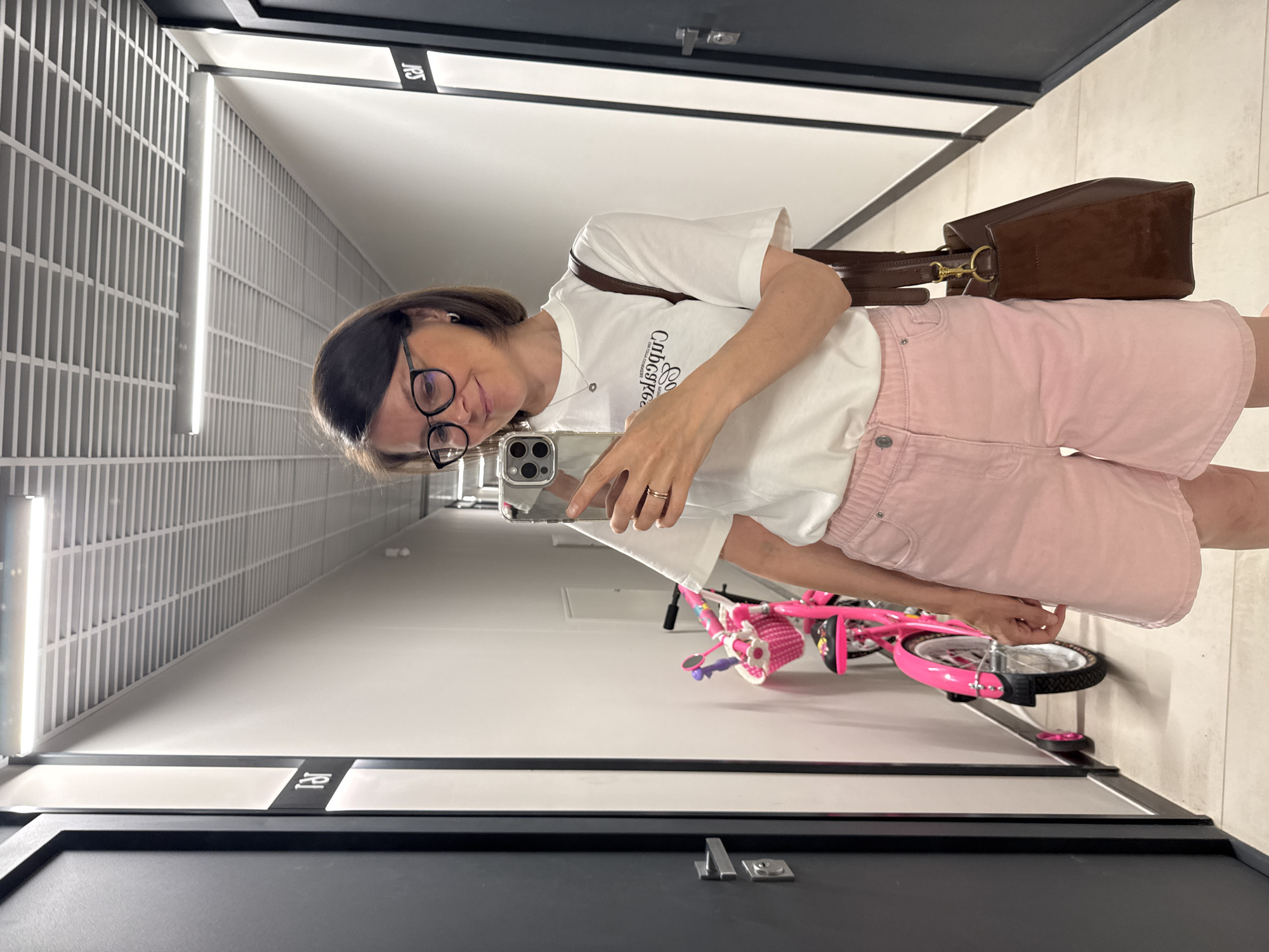 A woman stands in a hallway and takes a mirror selfie. She is wearing glasses, a white t-shirt, and pink shorts. She is holding a phone in her right hand and has a brown bag over her left shoulder. To her left is a pink bicycle. The hallway has white walls and a white tiled ceiling with fluorescent lights. Dark gray doors are visible on either side of the hallway. The woman has dark brown hair and is smiling. The scene appears to be in an apartment building or similar residential complex. Based on the provided information, the location is Izhevsk, Russia.