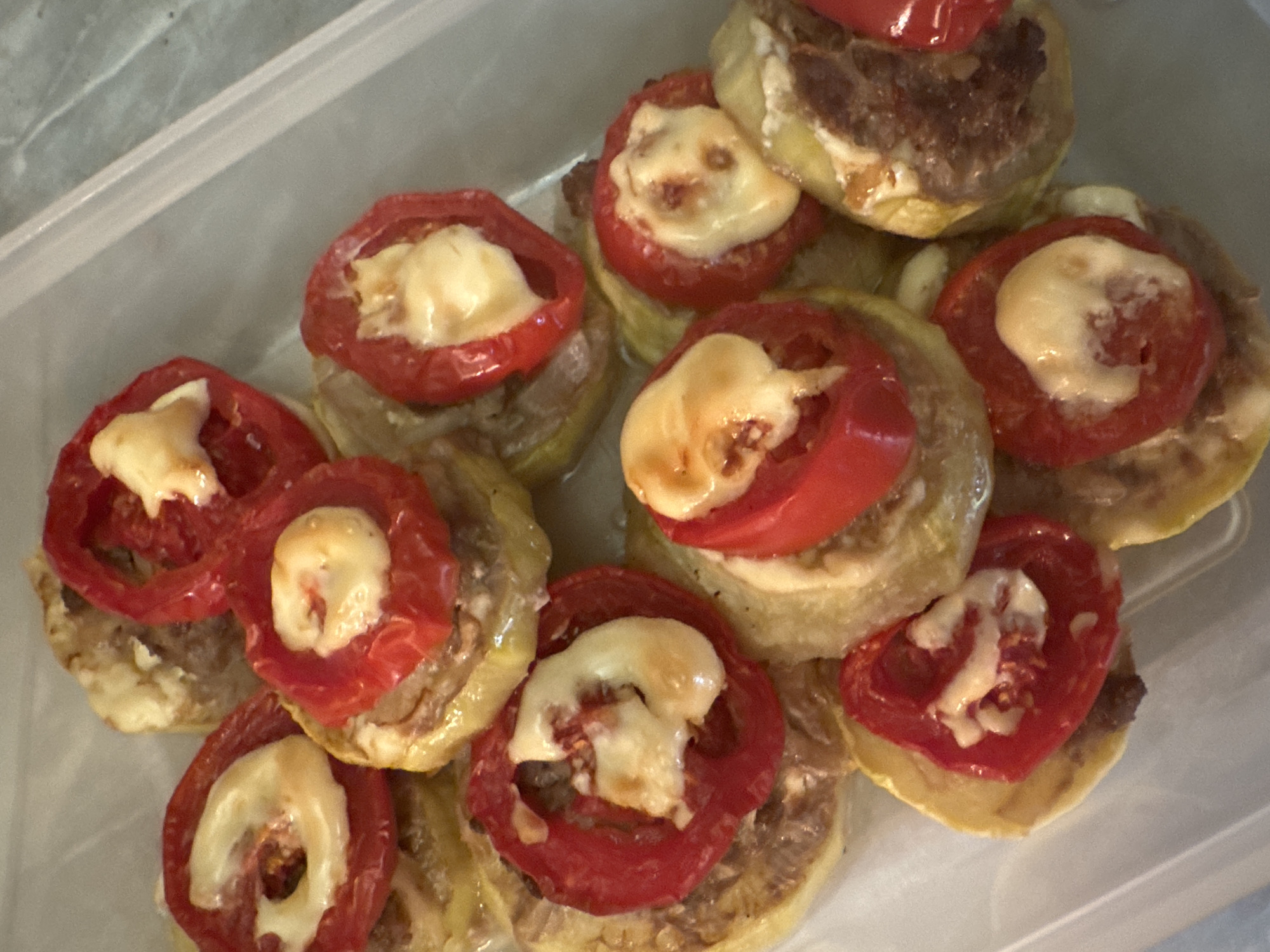The image displays a close-up, overhead view of a plastic container filled with baked zucchini rounds topped with tomato slices and melted cheese. The zucchini slices are golden brown and appear to be cooked through, with some browning around the edges. The tomato slices are bright red and have a slightly softened texture from baking, with melted white cheese in the center of each slice. The dish is densely packed within the container, with multiple layers visible. The container itself is clear plastic, with a textured surface on the right side, suggesting it might be a food storage container. The background is out of focus and appears to be a light-colored, possibly textured surface. There are no people or any specific city landmarks visible in the image. The scene is limited to the food item and its immediate container.