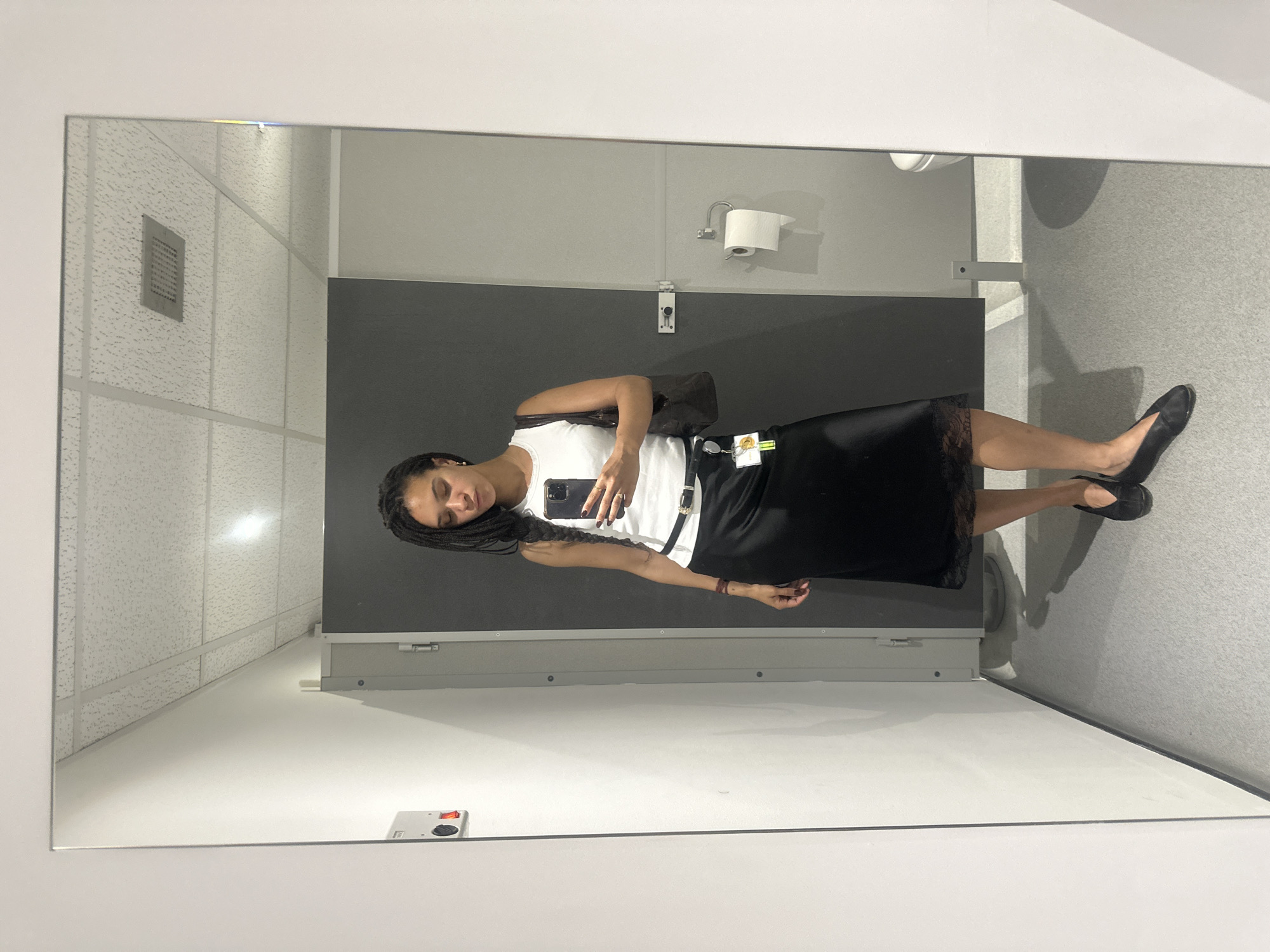 A woman stands in a bathroom stall, reflected in a full-length mirror. She has dark hair in long braids and wears a white tank top, a black skirt with lace trim, and black flats. She holds a smartphone in her hand. The bathroom stall has gray walls, a black door, and a white ceiling with recessed lighting. A toilet paper dispenser is visible on the wall. The location is a bathroom stall, likely within a public building in London, United Kingdom.