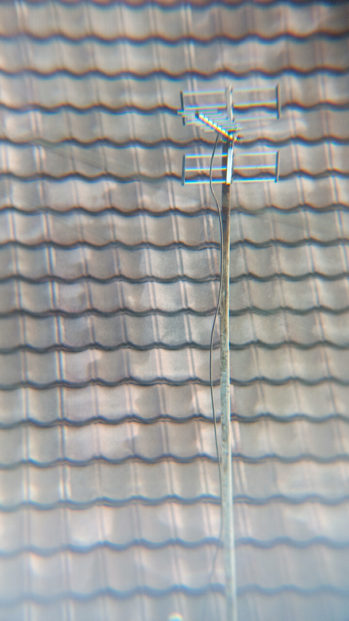 The image shows a television antenna mounted on a pole against a backdrop of tiled roofing. The antenna is positioned in the center of the frame and appears to be of a standard design with multiple horizontal elements. A black cable runs from the antenna down along the pole. The roofing tiles are arranged in horizontal rows, with a consistent wave pattern. The lighting suggests an outdoor daytime scene. The specific location of Margajaya, Indonesia, is not visually identifiable from the provided image.