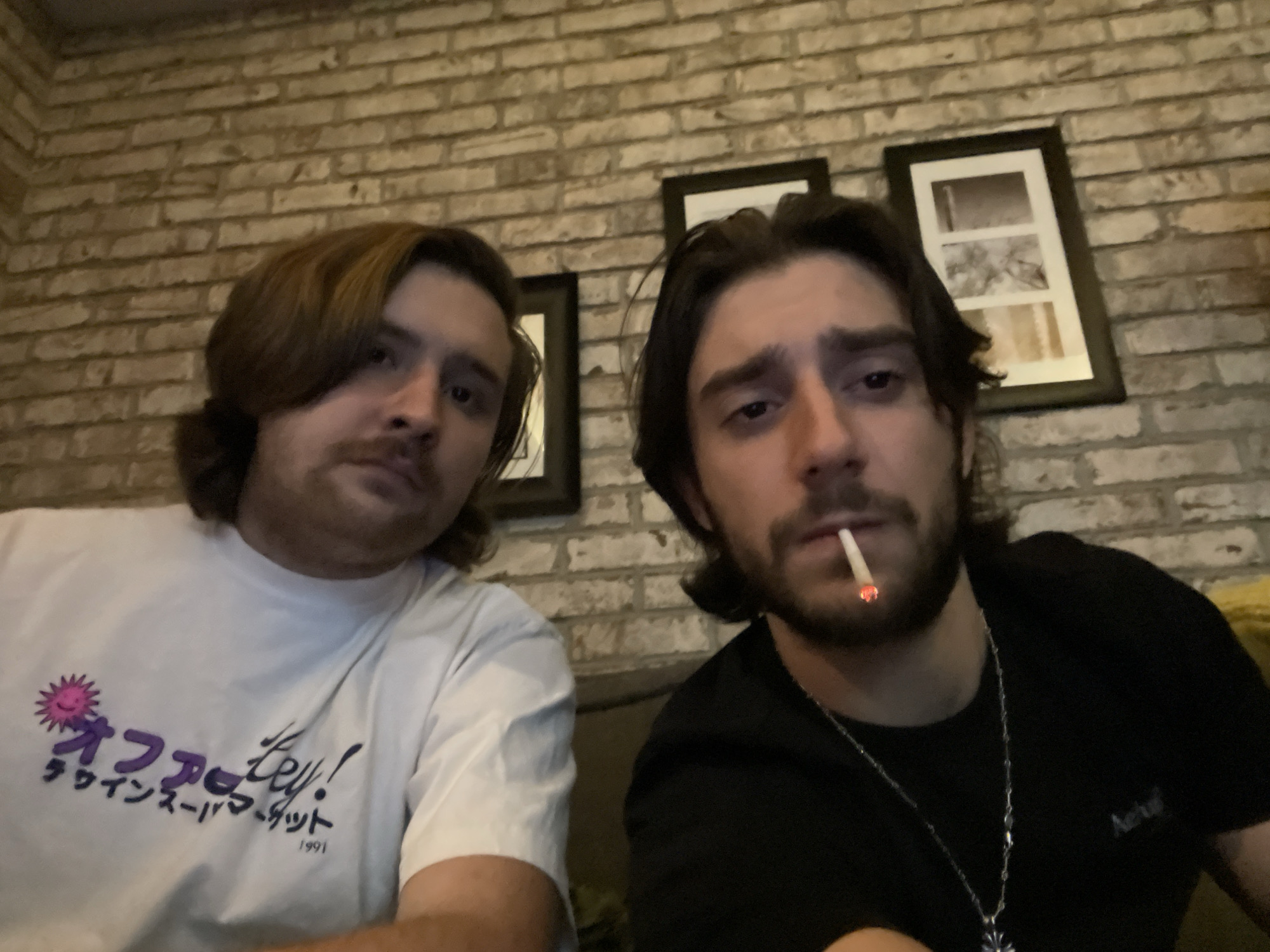 Two men are posing in front of a tan brick wall. The man on the left is wearing a white T-shirt with purple lettering. The man on the right is wearing a black T-shirt and a silver chain. He is holding a lit cigarette in his mouth. Two framed pictures hang on the wall behind them. The location is Huixquilucan de Degollado, Mexico.