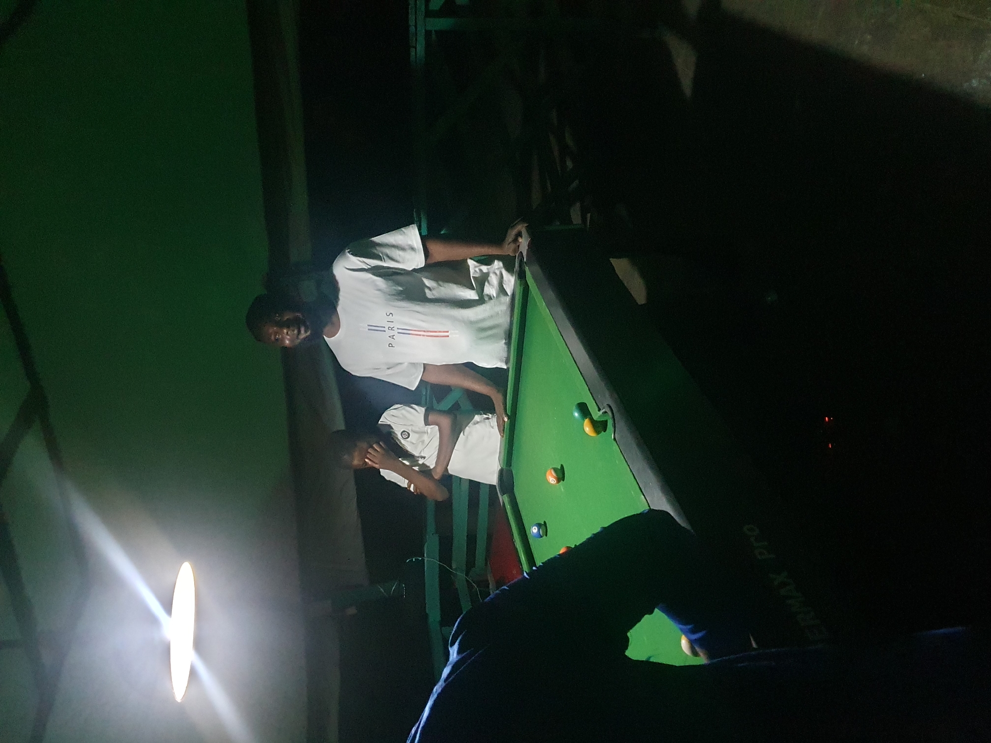 In Lokoja, Nigeria, two individuals are observed around a pool table under artificial lighting. One person, wearing a white t-shirt with "PARIS" printed on it, stands to the right of the table, looking forward. Another individual, partially obscured and wearing a white polo shirt, stands behind the table, with their hand near their face. A third person, mostly seen from the back and wearing a dark blue garment, is positioned to the left of the pool table, facing it. The pool table is covered in green felt and has several balls on it, including a blue ball, an orange and white striped ball, and a green and yellow striped ball. The environment appears to be an outdoor or semi-outdoor setting, possibly a recreational area, with a dark ceiling and green walls or partitions. The lighting is dim, with a bright, oval-shaped light fixture visible overhead. The overall scene suggests a casual game of pool in progress.
