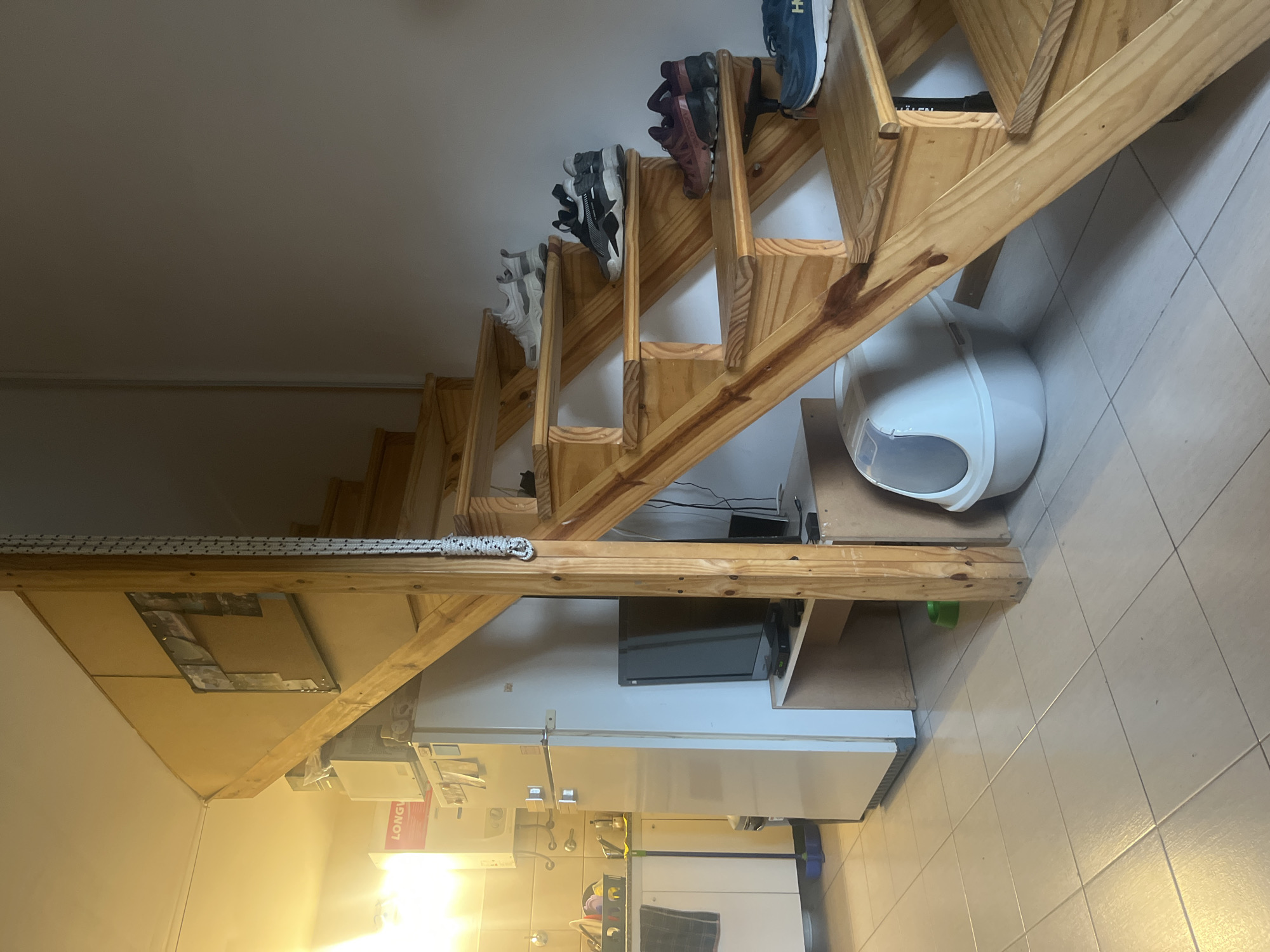 The photograph shows an interior space featuring a wooden staircase with shoes placed on the steps. A white refrigerator is visible below the staircase. There is an enclosed cat litter box next to the staircase. The lighting appears to be a mix of natural and artificial light. Details suggesting the location is Ushuaia, Argentina are absent.