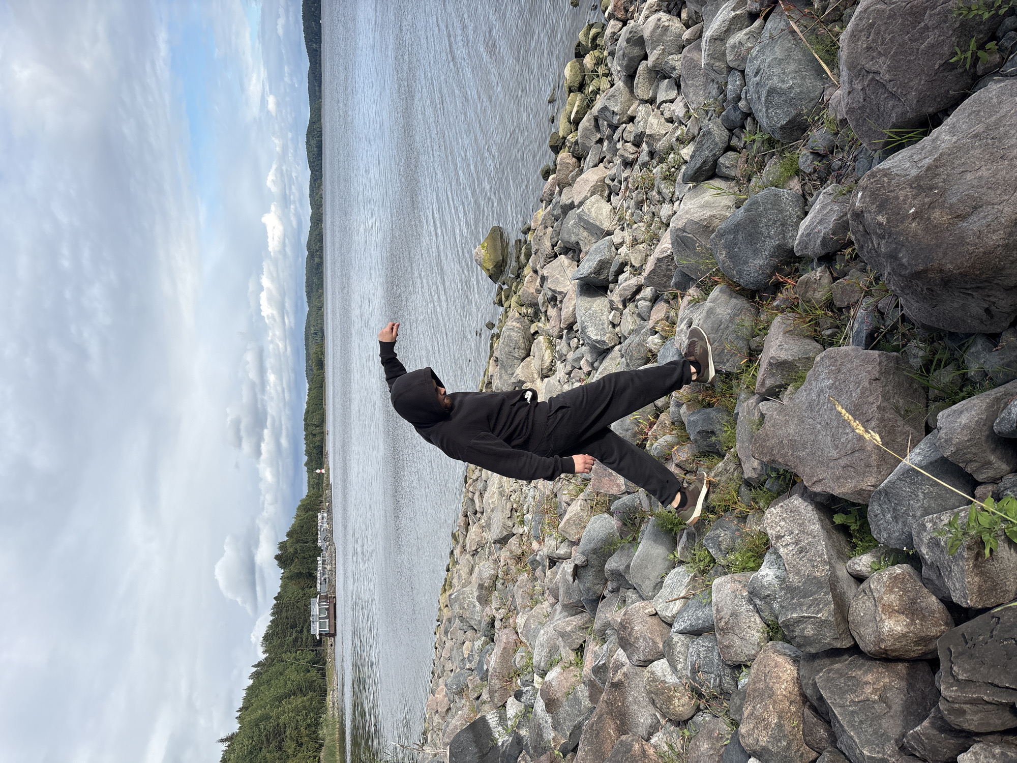 A person wearing a black hooded sweatshirt and black sweatpants walks across a rocky shoreline. The person's left leg is extended forward, and their right arm is raised and bent. The person is wearing brown shoes with white soles. The shoreline is covered in large, grey and brown rocks, with some green grass growing between them. In the background, there is a body of water, possibly a lake or sea, with a distant shoreline lined with trees. A few buildings and a lighthouse are visible on the far shoreline. The sky is cloudy, with patches of blue visible. The scene appears to be in a natural, outdoor setting. The location is Rautalakhti, Russia.