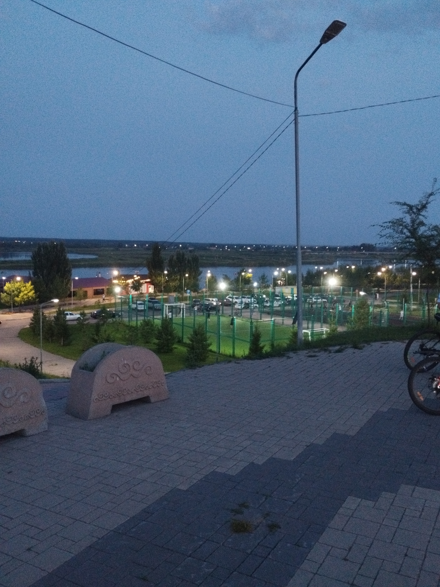 This image depicts an outdoor scene at dusk, possibly in Kostanay, Kazakhstan, given the location context. The sky is a deep blue, indicating the twilight hour. The foreground features a paved walkway with stone-like elements, possibly decorative benches or planters, on the left. In the middle ground, there's a sports complex with a green fenced area, likely a sports field or courts, illuminated by artificial lights. Several parked cars are visible near the sports complex, and a few people are seen walking around, suggesting the area is in use. Beyond the sports complex, a body of water, possibly a river or lake, stretches across the horizon, with lights from buildings and streetlights visible on the far bank. A tall lamppost stands prominently in the middle right of the image, casting light. On the far right, the rear wheel and frame of a bicycle are visible, suggesting someone has arrived by bike. The overall atmosphere seems peaceful and serene, as evening activities begin to wind down. The lighting conditions suggest it's either early morning or late evening.