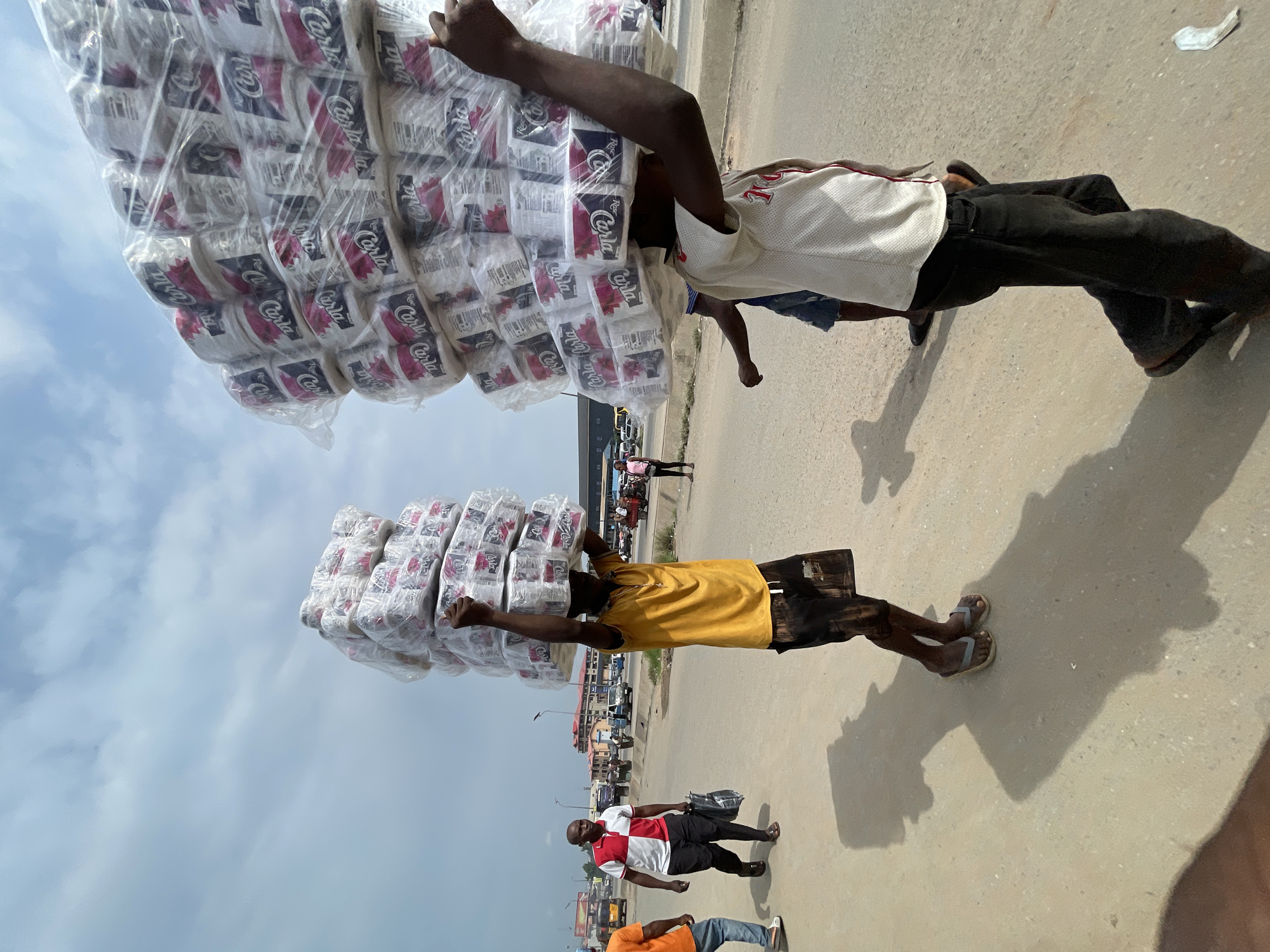 In a sunlit urban environment, two men are seen carrying large stacks of toilet paper rolls on their heads. The man in the foreground, wearing a white and red t-shirt, is facing away from the camera and appears to be walking to the right. The second man, to his left and closer to the camera, is wearing a yellow t-shirt and black shorts, also carrying a substantial load of toilet paper. Both men are on a paved road with visible tire tracks, and shadows are cast on the ground. In the background, other individuals, vehicles, and buildings suggest a bustling marketplace or street scene. The sky above is partly cloudy.