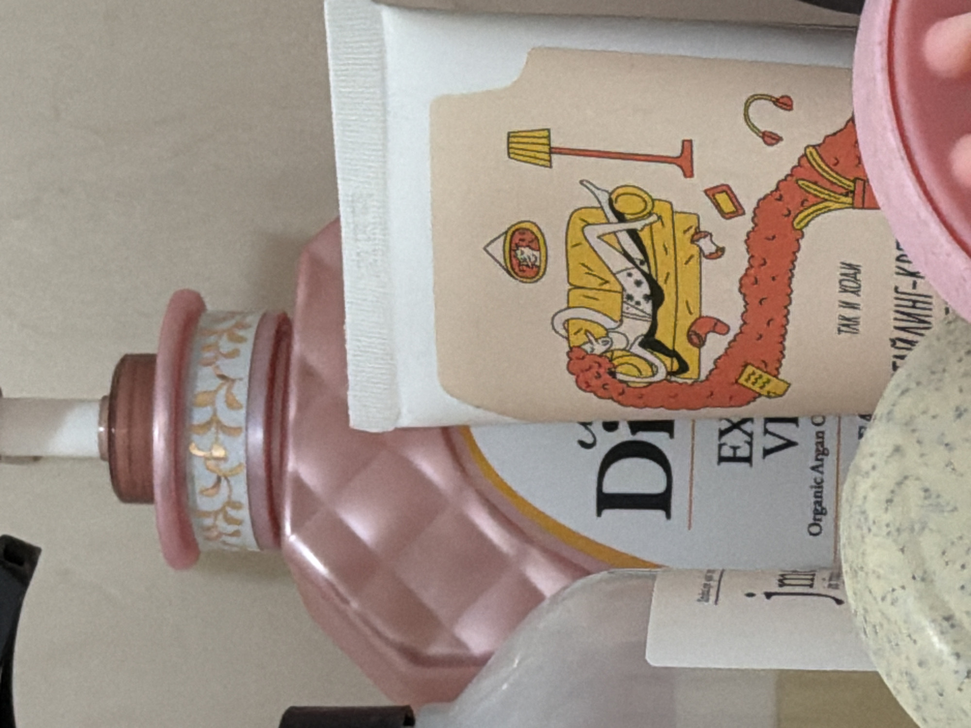 The image shows a close-up of several beauty products. In the foreground, a light-colored, textured object, possibly a bath bomb or exfoliating scrub, is partially visible. Behind it, a pink bottle with a diamond-patterned texture and gold floral accents is prominent. To the right of the pink bottle is a white tube with a cartoon illustration of a person lounging on a yellow couch, with a red wavy shape flowing from the couch and a yellow lamp standing nearby. Text in Cyrillic script is visible on both the pink bottle and the white tube. The background is a plain, neutral surface, suggesting an indoor setting like a bathroom or vanity. There are no people or animals visible in the image, nor are there any discernible landmarks or environmental features that would indicate a specific location in Moscow, Russia.
