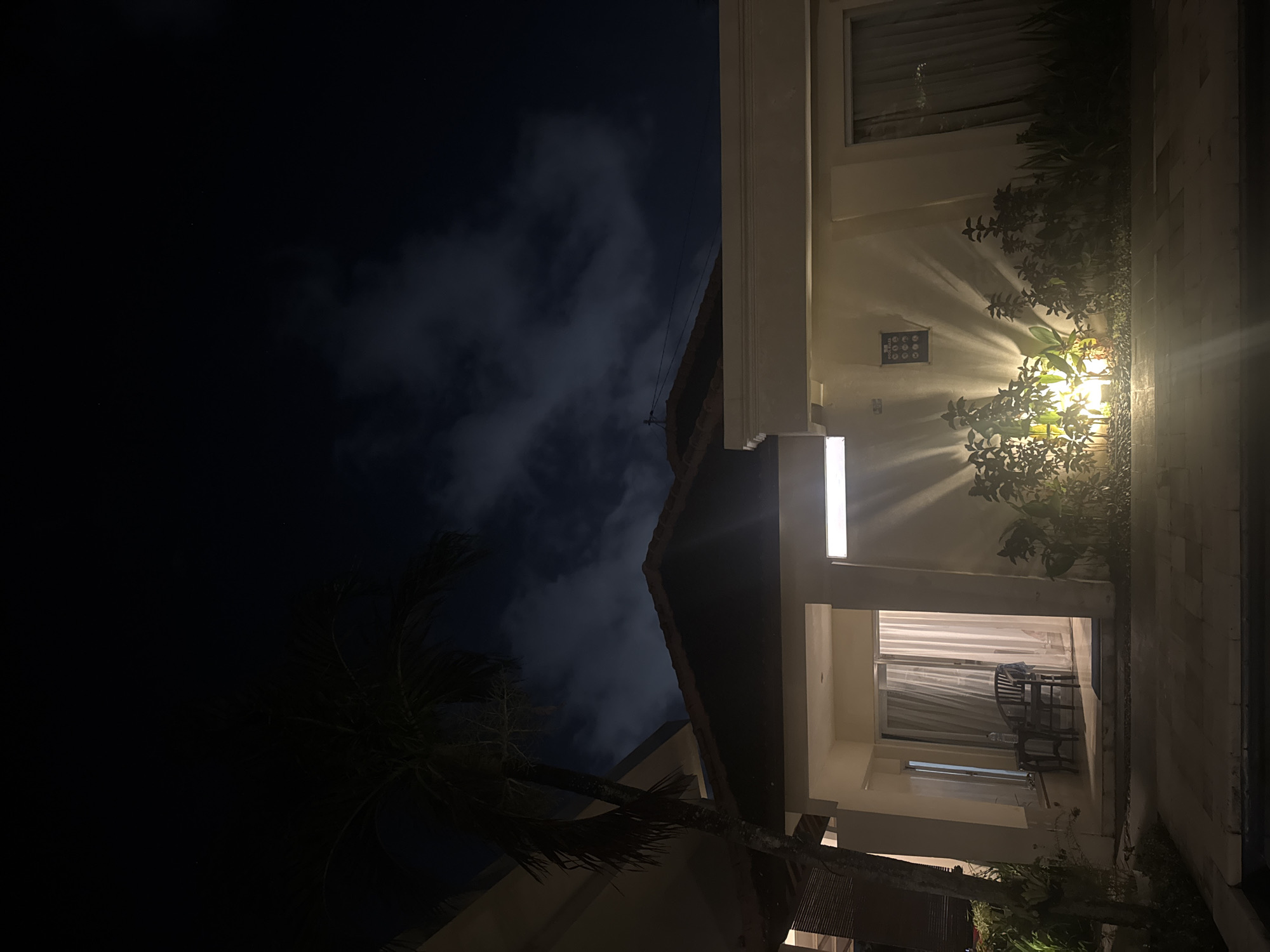 This media file depicts a nighttime scene outside a building, likely a hotel or resort, in Kuta Selatan, Indonesia. The image is oriented vertically, with the sky occupying the left portion and the building's exterior on the right.
The sky is dark, with wispy clouds visible, suggesting a partly cloudy night. A silhouette of a palm tree frond is present in the lower left, hinting at a tropical location. The building features a modern design with white and beige elements. A warm-toned outdoor light illuminates a patch of greenery and the facade of the building, casting dramatic shadows. The light appears to be a wall-mounted fixture shining downwards.
A small, square blue plaque with a white grid of nine circular symbols is mounted on the wall near the light. This could be a warning sign or informational plaque. Further along the building, an open doorway reveals a well-lit room with sheer curtains covering a window. Inside the room, two dark wooden chairs and a small table are visible.
The overall mood is calm and serene, amplified by the warm lighting and the quiet of the night. The time of day is clearly night. The location cues, such as the palm tree and the architectural style, are consistent with a tropical resort setting in Indonesia. No discernible text is present on the plaque, other than the symbolic representations.