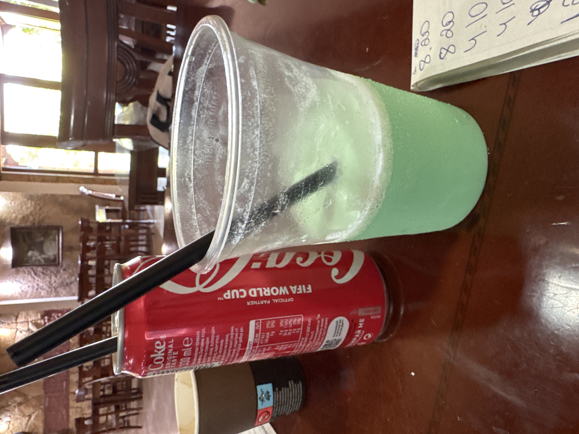 A close-up, eye-level, outdoor shot shows two black straws in a can of Coca-Cola on a wooden table. To the right of the Coca-Cola can is a clear plastic cup with a green beverage, and a small notebook with handwritten numbers. The background shows a restaurant interior with tables, chairs, and a window. The Coca-Cola can is a standard 330ml size and is labeled "FIFA World Cup." The green beverage in the plastic cup appears to be a slushy or frozen drink. The notebook has entries such as "8.20", "4.10", and other illegible writing. The overall lighting suggests a bright, daytime setting. The presence of the Coca-Cola can and the restaurant setting suggest a casual dining or refreshment experience. This scene could be found in any establishment serving beverages, including those in Sredets, Bulgaria.
