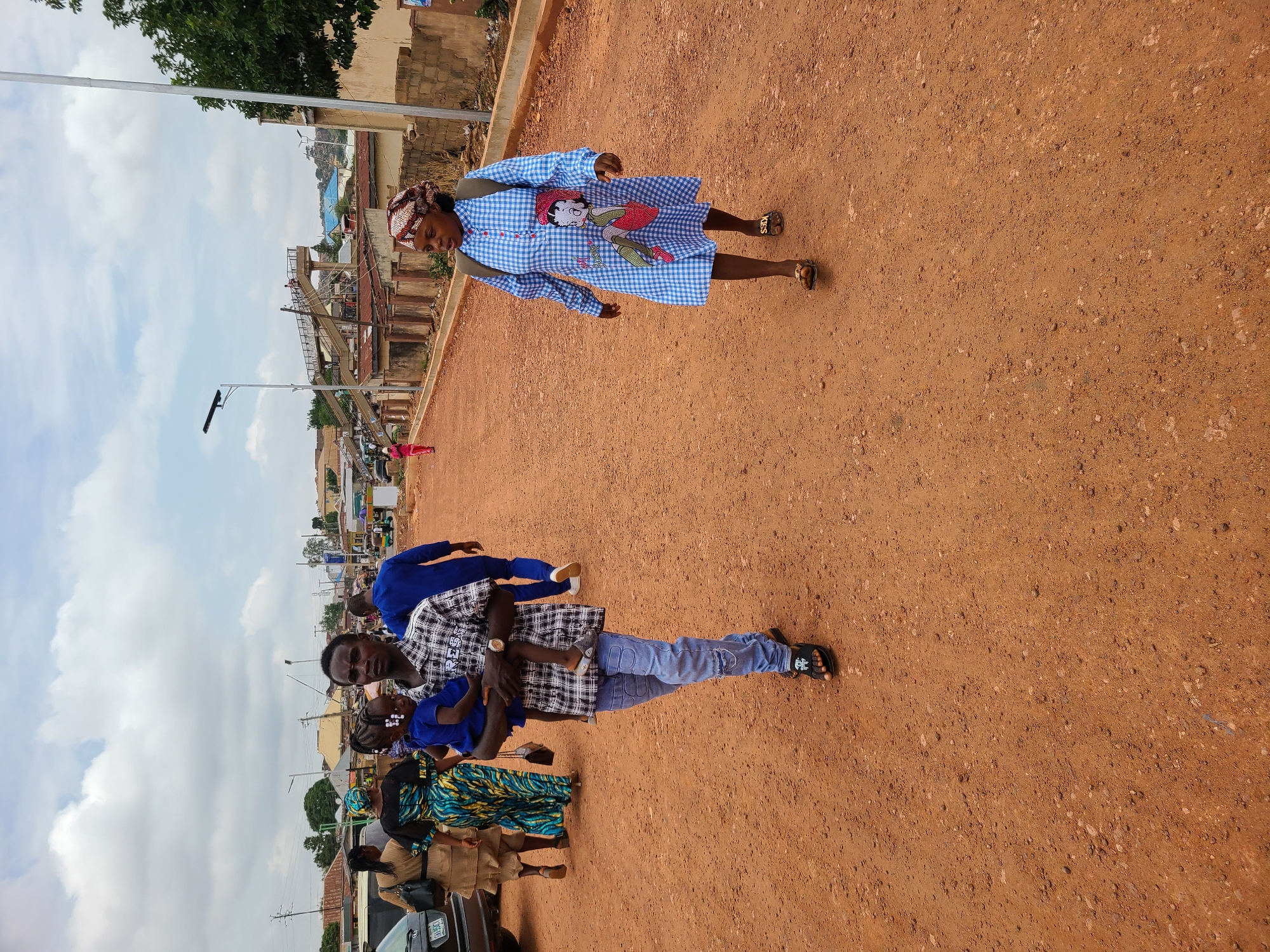 The image shows a group of people standing on a reddish-brown, dirt or gravel ground in an urban environment.
A woman is wearing a blue and white checkered dress with a cartoon character on it. She has a handbag slung over her shoulder.
A man is holding a child, wearing a blue shirt and jeans.
Other people are nearby, some in what appear to be school uniforms with a patterned skirt.
Behind the people are cars and buildings. There is what looks like a construction site for a bridge or elevated road. Street lights are also visible.
The location appears to be outdoors during the day, with a cloudy sky.