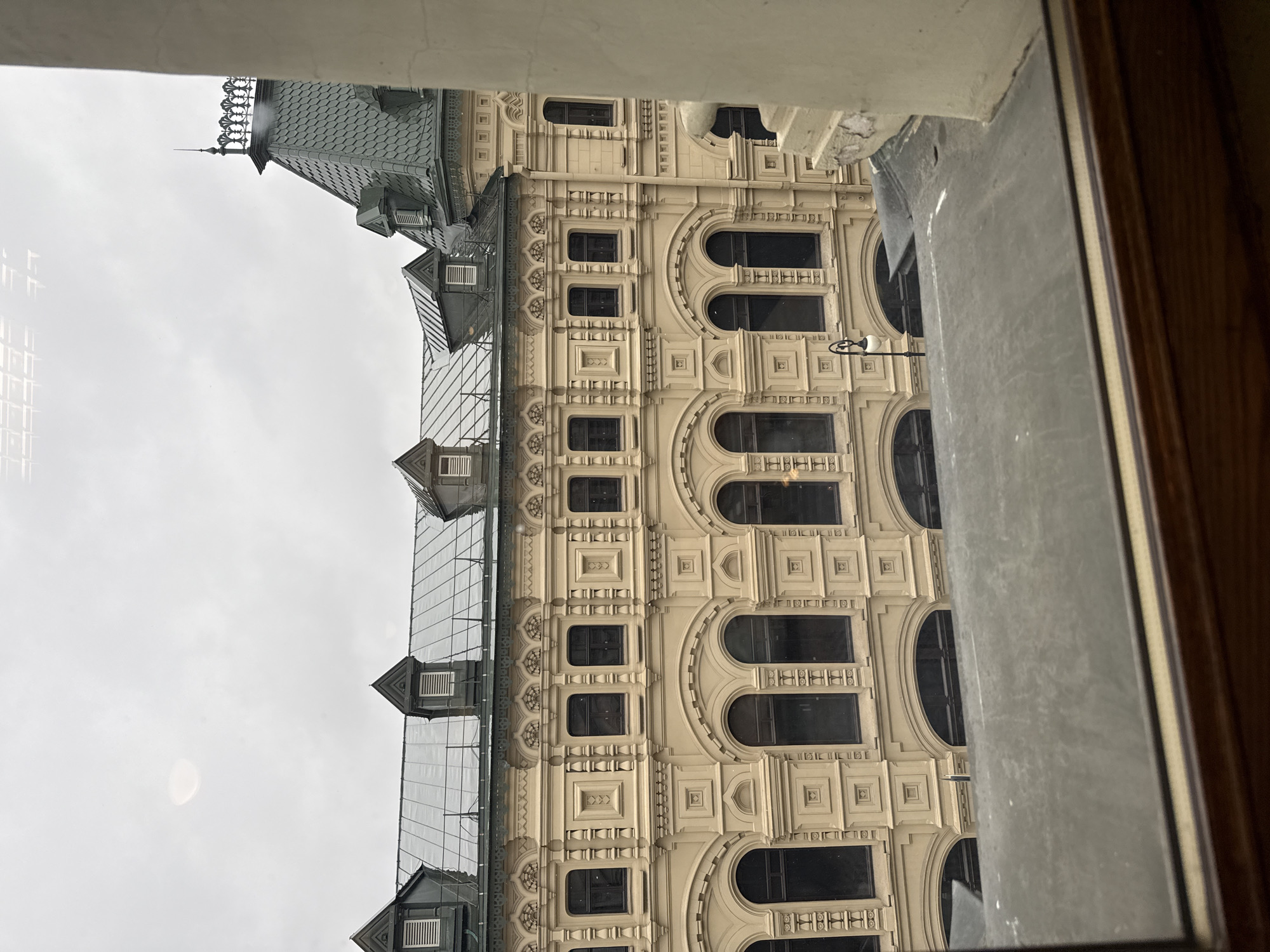 From a window perspective, the image captures the exterior facade of the GUM department store in Moscow, Russia. The building features a symmetrical design with multiple floors, ornate detailing, arched windows, and a glass roof. The sky is overcast. A portion of the window frame is visible in the foreground.