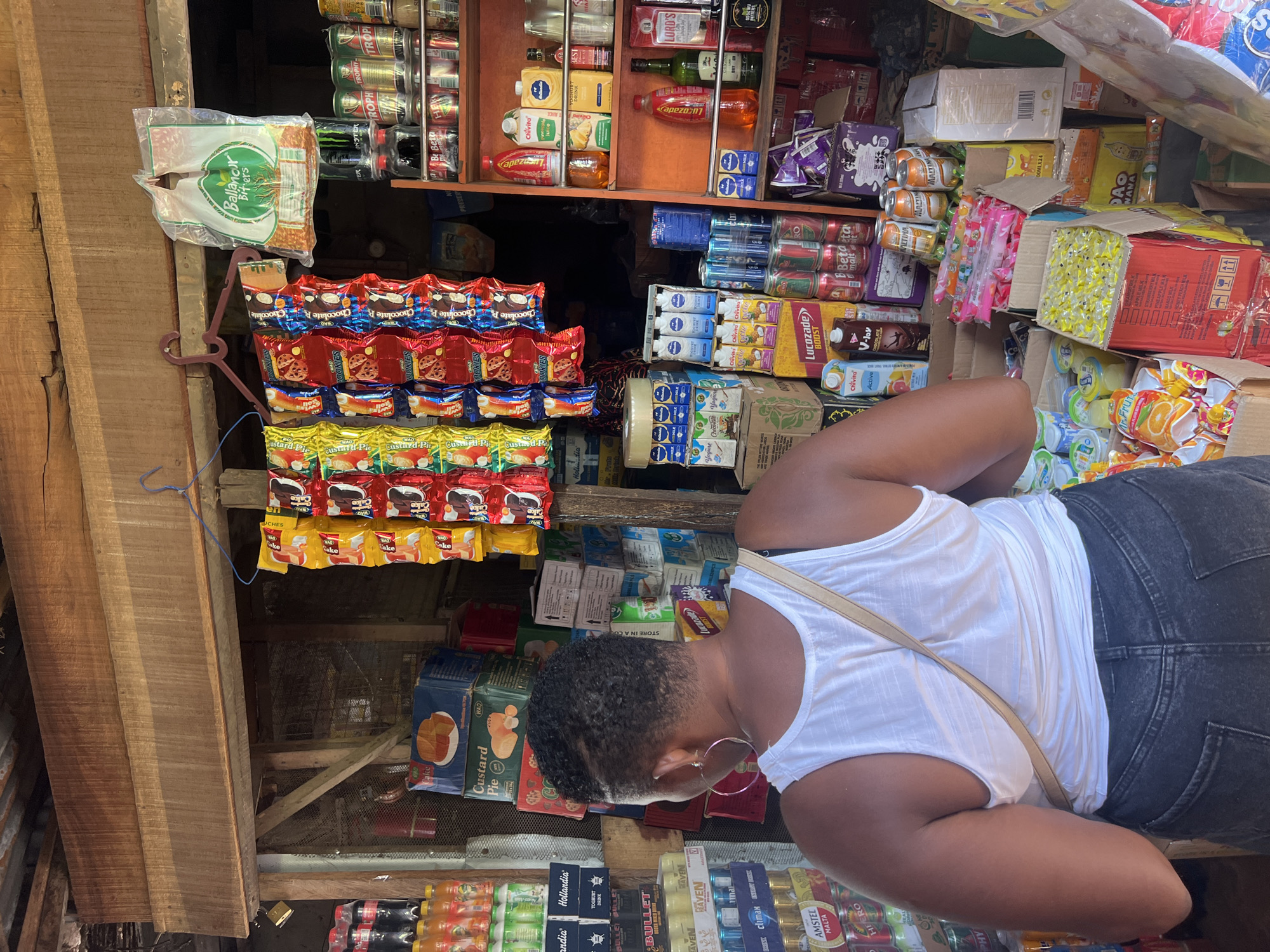 A woman with short dark hair, wearing a white tank top and dark jeans, stands examining a shop stall in Nkpor, Nigeria. She wears hoop earrings and has a purse strap across her chest. The shop is stocked with a variety of goods, including packaged cookies, custard pie mix, yogurt drinks, canned beverages, and bottles of various liquids. A sign reading "Ballanour Biters" is visible. The shop is constructed of wood and metal, with goods displayed on shelves and hung from hooks.