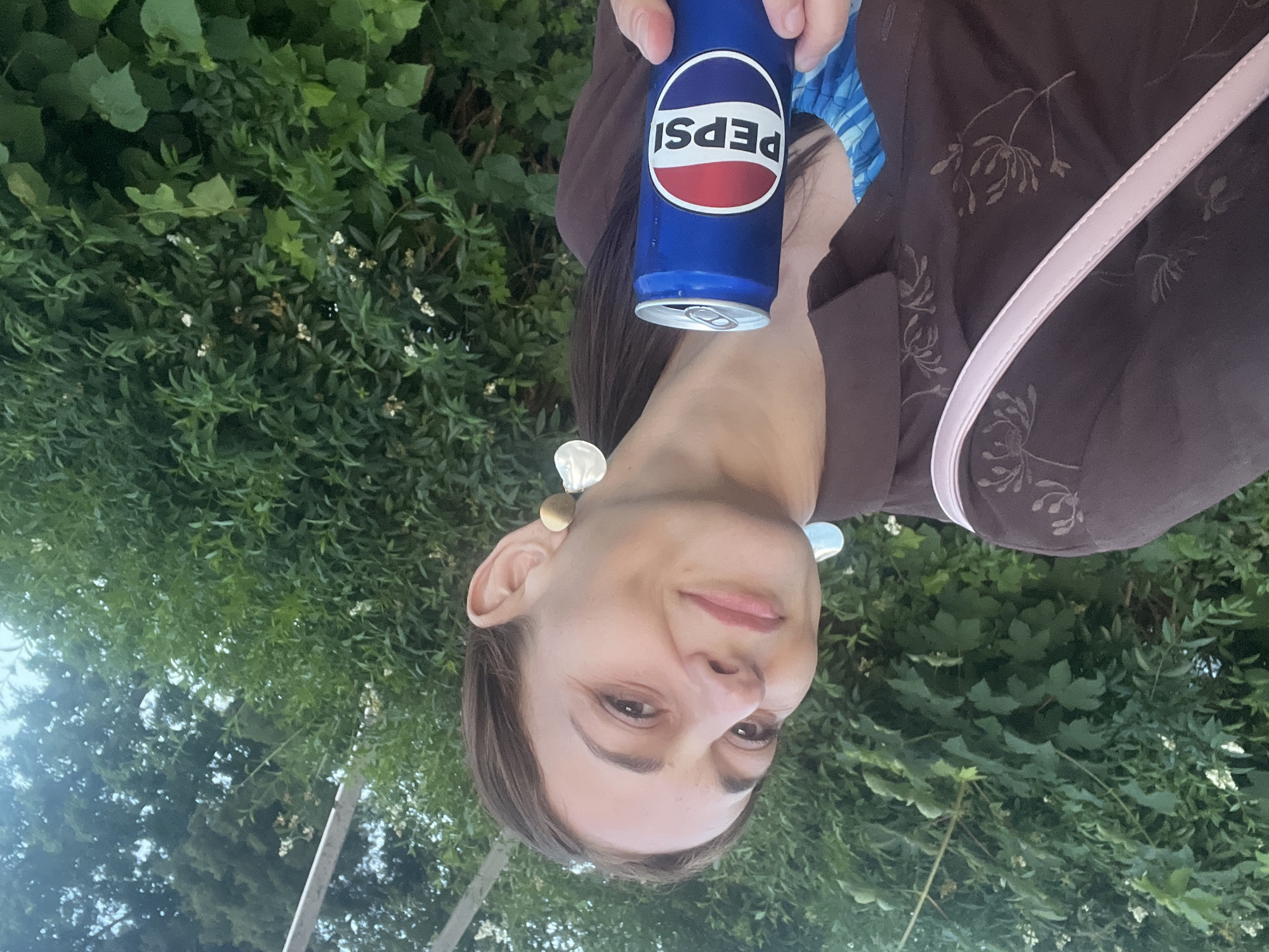 A woman is holding a can of Pepsi while standing outdoors, with green foliage behind her. She is wearing a brown shirt with a floral pattern and a pink strap across her shoulder. The Pepsi can is blue with the iconic red and white logo. The woman is looking at the camera and smiling. The background suggests a natural setting, possibly a park or a garden.