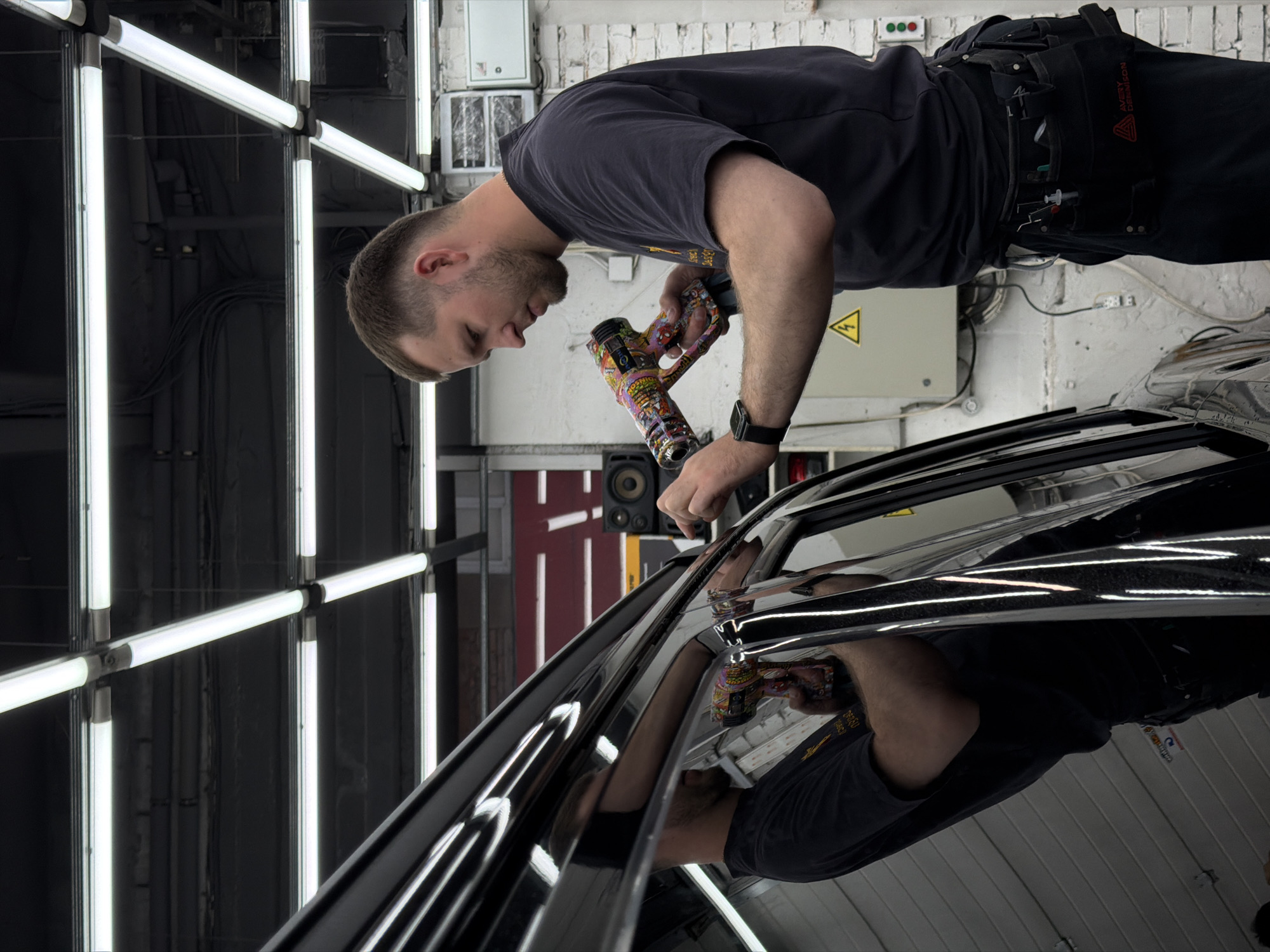 A man with short brown hair, wearing a dark T-shirt and dark pants with a tool belt, is working on a black car. He is holding a tool with a colorful design on it. The car's surface is shiny and reflective. The surroundings suggest an automotive shop or garage, with a white brick wall in the background, fluorescent lights on the left, and various electrical boxes and equipment visible. The location may be Krasnoyarsk, Russia.