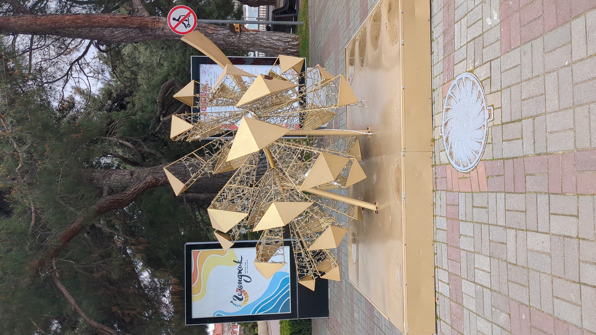 The image shows a golden, geometrically-shaped art installation on a brick-paved sidewalk in Gelendzhik, Russia. The installation appears to be a modern sculpture featuring a central hub with triangular and cage-like elements radiating outward. It is mounted on a rectangular, gold-colored base. Adjacent to the installation is a poster with the word "Геленджик" (Gelendzhik) written in Russian script, along with a colorful, wavy design suggesting a seaside theme. A sign prohibiting some activity (possibly walking on the trees) is visible on a tree branch in the background. A utility hole cover is visible on the ground.