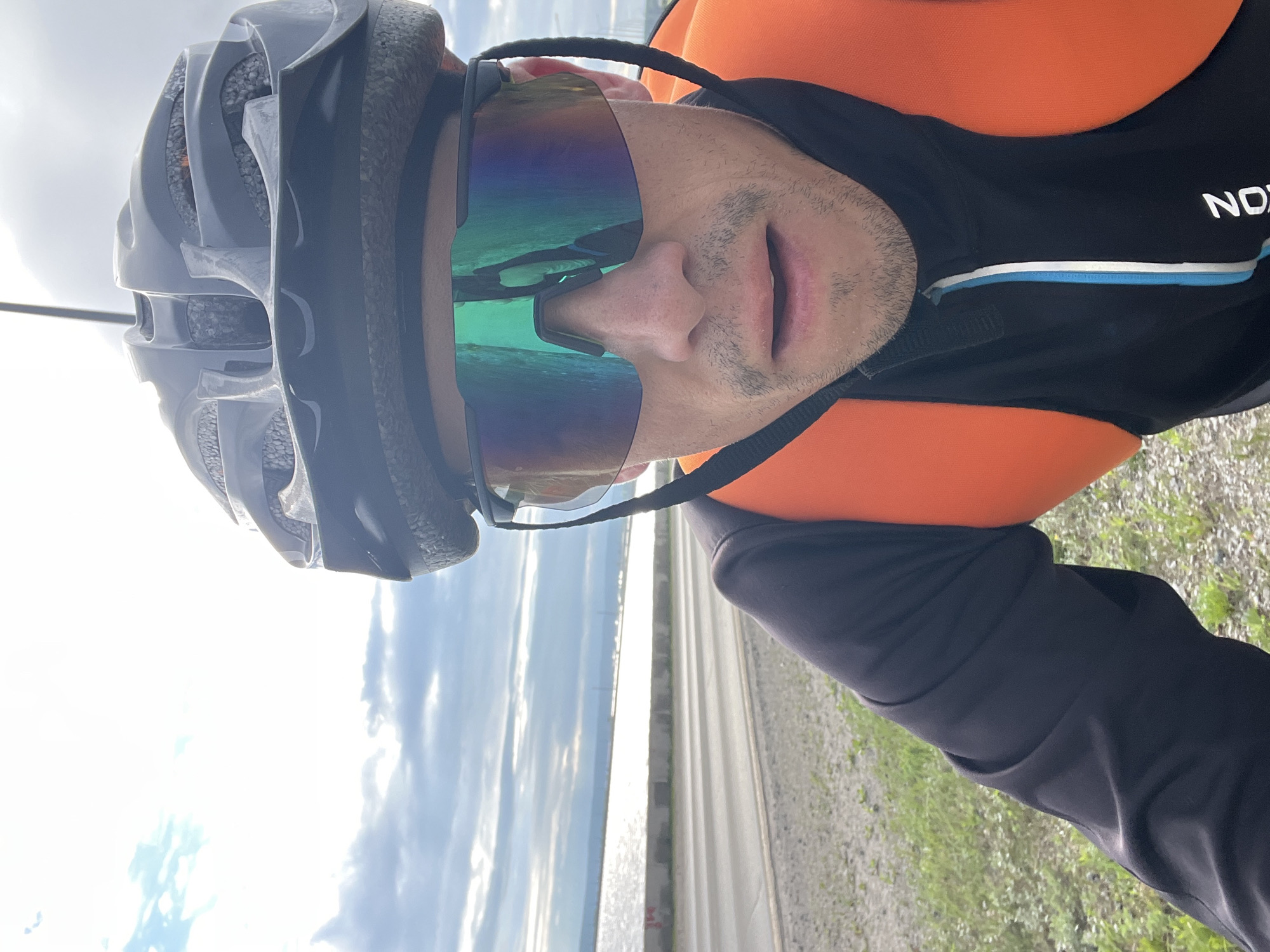 A man wearing a black helmet, cycling sunglasses with a green-blue tint, a black cycling jersey with an orange vest, and a backpack is taking a selfie while cycling outdoors. The man has a light beard and is looking forward with his mouth slightly open. In the background, there is a road, a body of water that appears to be the Kama River, and a cloudy sky. The scene suggests an outdoor activity, possibly training or recreation, in a setting that could be along a coastal or riverside path. The provided coordinates are for Chaikovsky, Russia, suggesting this is where the photograph was taken.