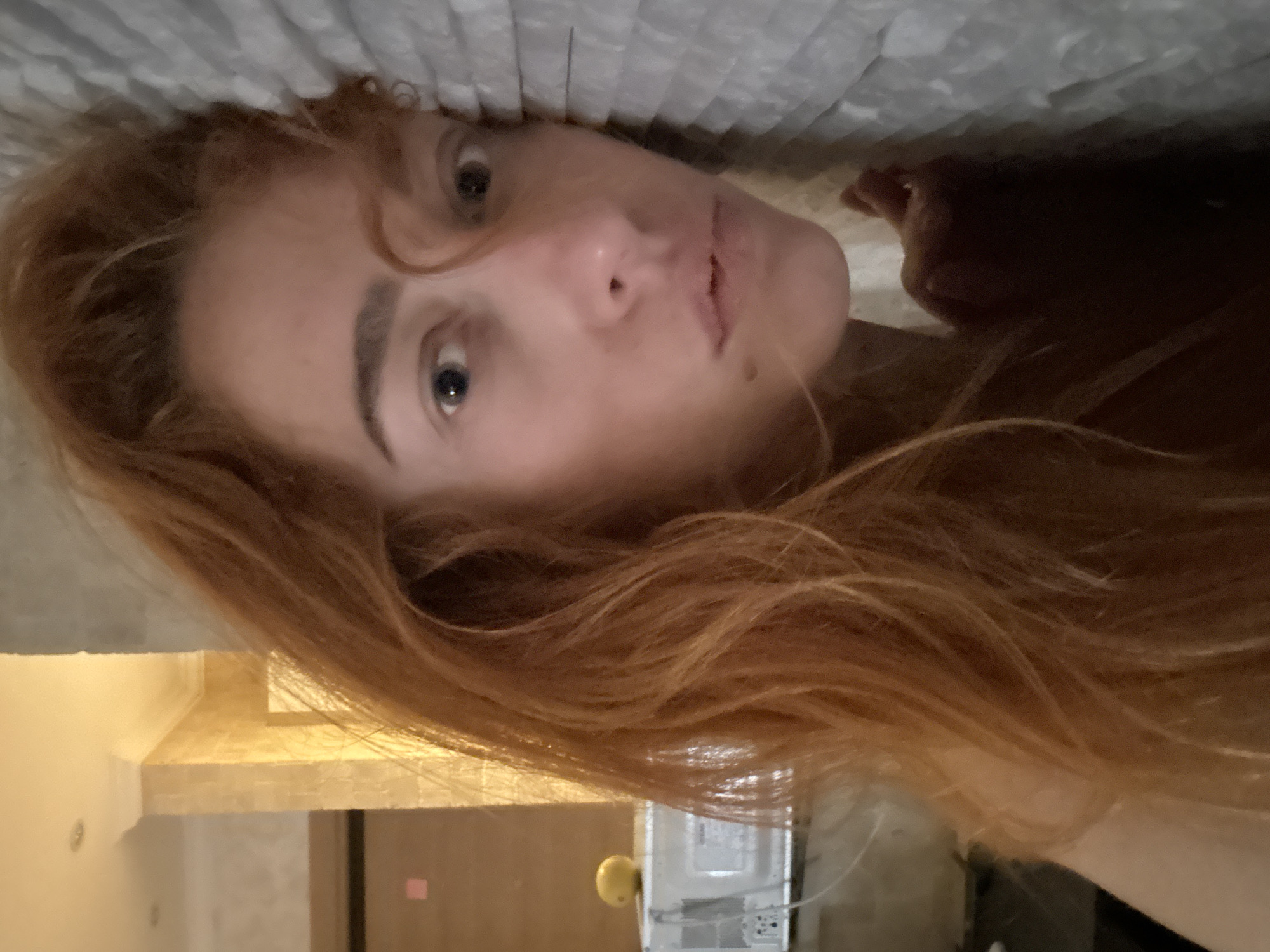 A woman with red hair is lying on a bed with a white comforter, appearing to be indoors. Her face is the primary focus of the image. The background shows a room interior, possibly a hotel room, with what appears to be a door and a side table with an object. Given the reference to Budva, Montenegro, it can be inferred that the scene is taking place in a hotel or residence within the city.