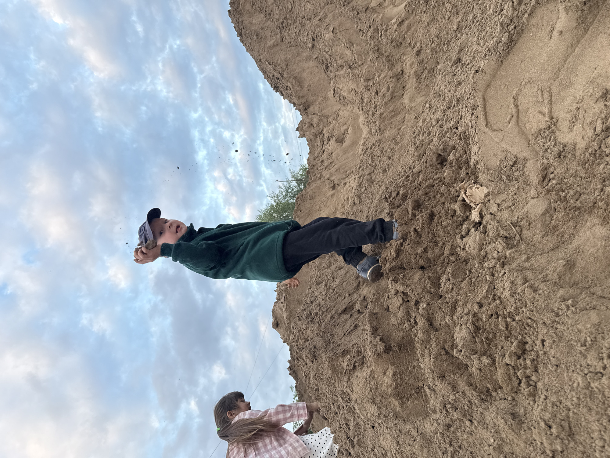 In the city of Chaikovsky, Russia, two children are playing on a large pile of sand under a cloudy sky. A boy, wearing a dark green jacket and a blue baseball cap, is in the foreground, throwing a handful of dirt into the air. He is on top of the sand pile, facing the camera with a playful expression. To his left, a girl with long brown hair tied in a ponytail is partially visible, also on the sand pile, wearing a pink and white checkered shirt and a white skirt with polka dots. The sand pile dominates the scene, with the sky filled with white and gray clouds providing the backdrop.