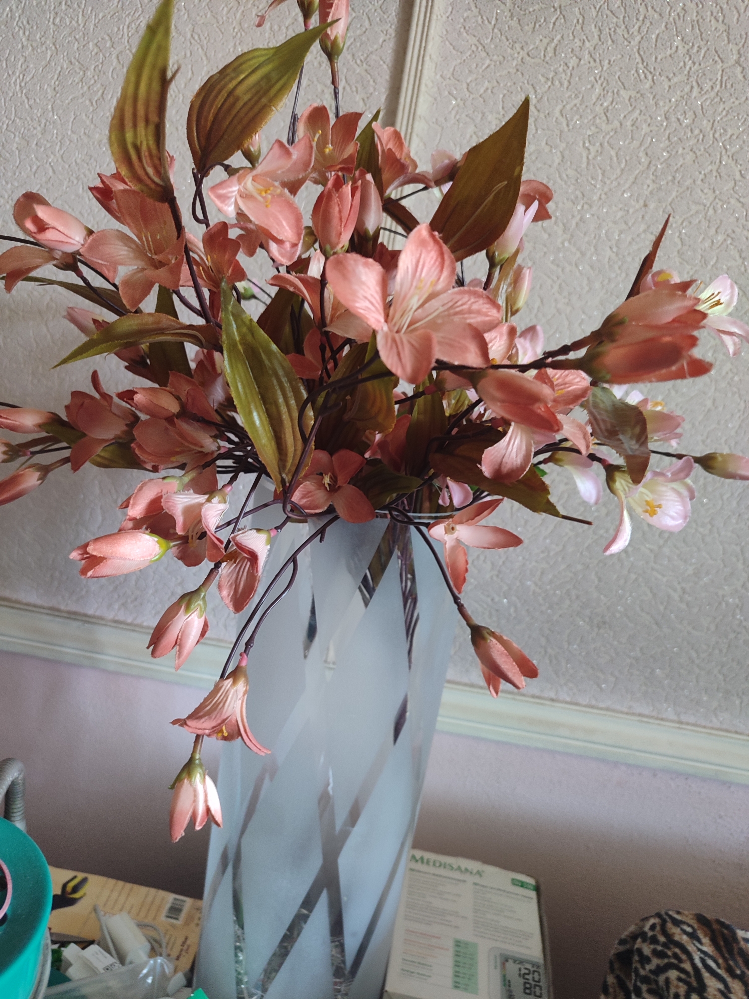 An artificial floral arrangement of pink lilies and green foliage is displayed in a tall, frosted glass vase with a patterned design. The vase is positioned on a surface, with a ceiling visible in the upper portion of the image. The background consists of textured wallpaper and a white molding. Below the vase, a portion of a box for a "MEDISANA" blood pressure monitor and a leopard-print fabric are visible. The scene appears to be indoors.