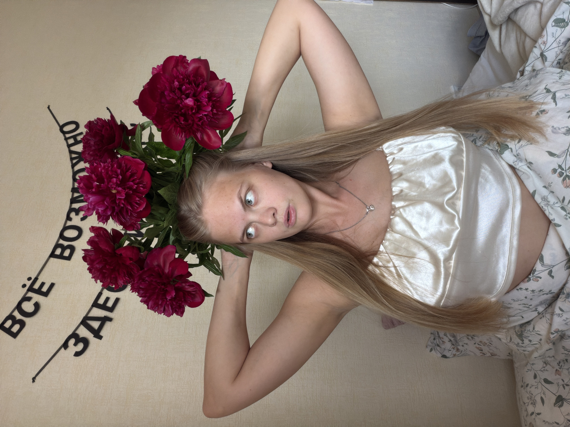 A high-angle, portrait-oriented photograph (rotated counter-clockwise) depicts a young woman lying down in an indoor setting. She has long, straight blonde hair and fair skin, wearing a shiny, light-colored, possibly cream or white, satin crop top and a delicate silver necklace with a heart-shaped pendant. Her arms are raised and bent, with her hands positioned near her head.
A vibrant arrangement of large, deep red or burgundy peonies with green foliage is placed around her head, forming a striking visual similar to a floral crown or halo. The woman's eyes are wide open and directed towards the left side of the frame (her left), conveying an expression of surprise, curiosity, or perhaps playful wonder. Her lips are slightly parted.
She is situated on a light-colored, possibly beige, padded surface, which could be a bed or sofa. On the right side of the frame, a section of white bedding with a subtle green and light pink floral pattern is visible.
Behind her head, on the light-colored background, black Cyrillic text is prominently displayed. The visible words are "ВСЁ ВОЗМОЖНО ЗДЕСЬ," which translates to "EVERYTHING IS POSSIBLE HERE." The indoor setting, likely a domestic space, appears naturally lit, suggesting daytime. The presence of Cyrillic text aligns with the given location context of Izhevsk, Russia.