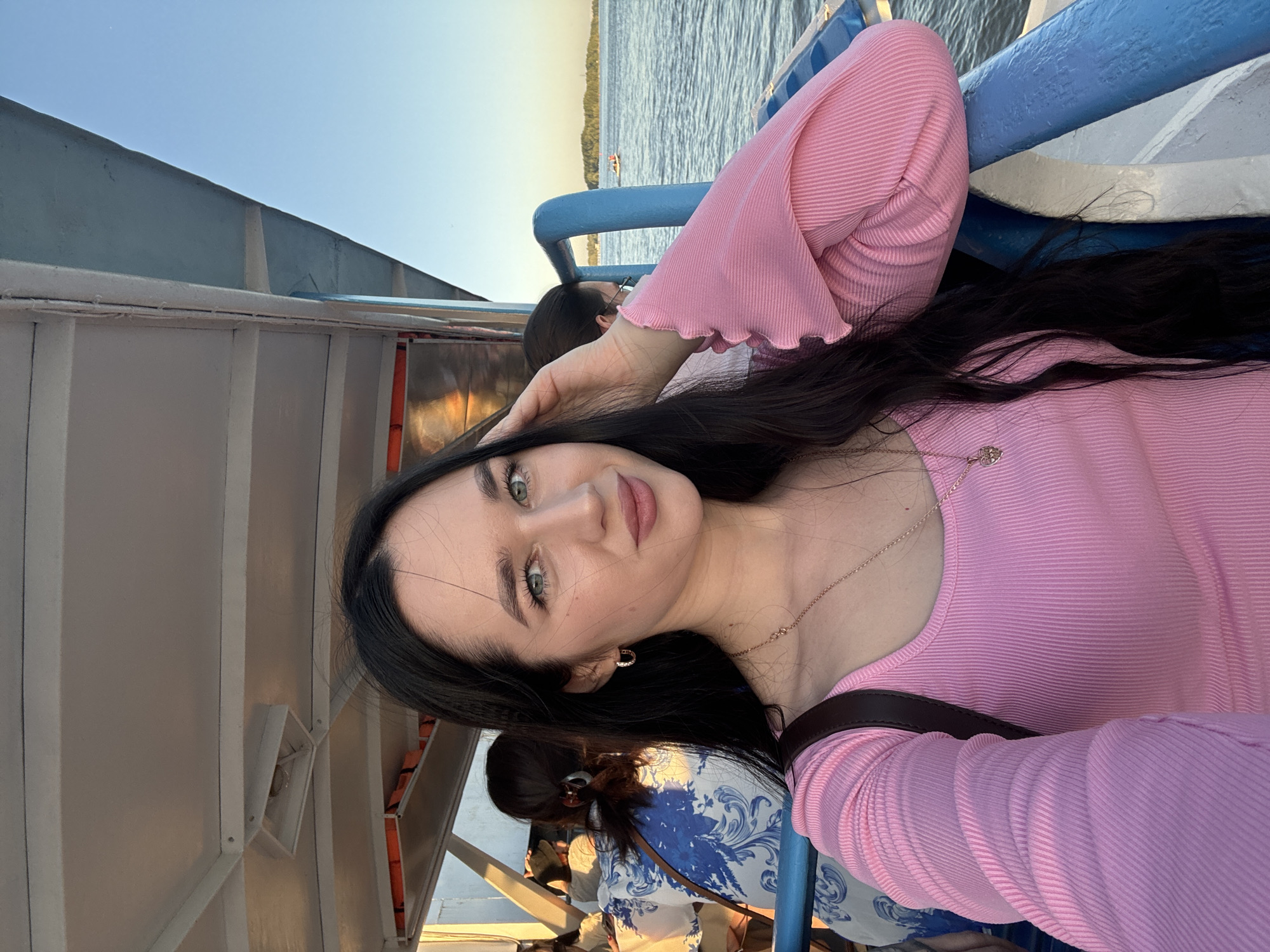 A young woman with long, dark hair and striking green eyes takes a selfie on a boat. She is wearing a pink, ribbed long-sleeved top with ruffled cuffs and a delicate silver necklace. Her left hand is resting on her head, and she is looking directly at the camera with a slight smile.
The setting appears to be a sunny day, likely late afternoon or early evening, given the warm light casting shadows. She is on a boat, with a blue railing visible in the foreground and the calm water of a river in the background. Other passengers are visible in the background, some seated and some standing. The boat has white siding and a pale blue railing. The sky is clear and light blue.
The image conveys a relaxed and joyful mood. The woman’s pose and smile suggest she is enjoying her time on the water. The light and clear sky indicate pleasant weather, perfect for a boat trip. The presence of other people suggests a public ferry or tour boat. The location context of Novosibirsk, Russia, implies the boat is likely on the Ob River. There is no visible text in the image.
