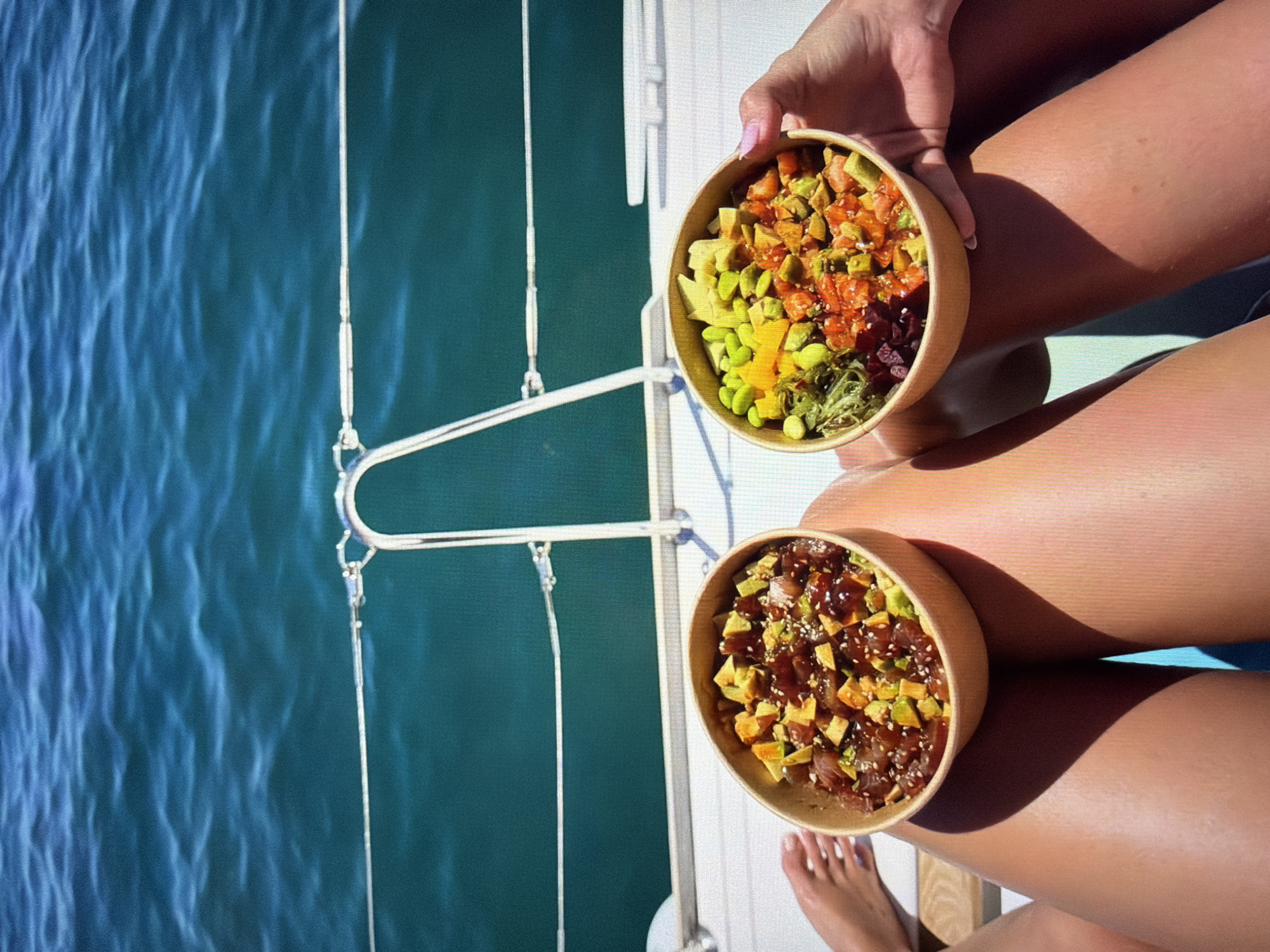 This vertically oriented image captures a serene scene aboard a boat or yacht near Ros Na Rí, Ireland. Two individuals are depicted, primarily by their tanned legs and hands, enjoying poke bowls.
In the upper right, a hand with pink nail polish holds a colorful poke bowl. This bowl contains a vibrant medley of diced salmon, avocado, edamame, bright yellow mango, and green seaweed salad, sprinkled with sesame seeds. Below it, another poke bowl is visible, resting near a second pair of tanned legs. This bowl features diced tuna, avocado, and a dark sauce, also garnished with sesame seeds.
The setting appears to be a sunny day, with clear sunlight casting distinct shadows on the legs, indicating warm and pleasant weather. To the left of the frame, the deep blue-green waters of the sea are visible, showing gentle ripples. Elements of the boat's rigging, including ropes and metal structures, along with the white deck, are present, confirming the maritime location. The overall scene suggests a relaxed and enjoyable meal on the water. There is no visible text in the image.