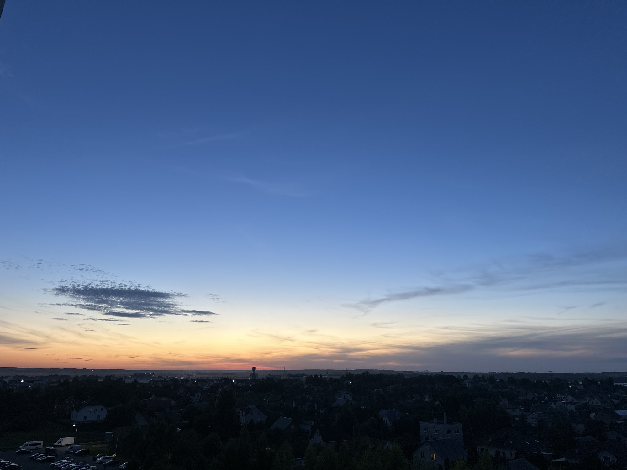 This is a wide-angle, high-angle shot of a suburban landscape at dusk, likely in the vicinity of Шчомысліцкі сельскі Савет, Belarus. The sky is a gradient of deep blue at the top transitioning to lighter blues and then to hues of orange, yellow, and pink near the horizon, indicating either sunrise or sunset. Wispy clouds are scattered across the sky, catching the light. Below the sky lies a dark silhouette of a residential area, characterized by numerous houses with visible roofs and trees. In the foreground, on the left side of the image, is a paved parking lot filled with several parked cars and a few white vans. The overall scene captures a quiet, evening atmosphere in a residential community.