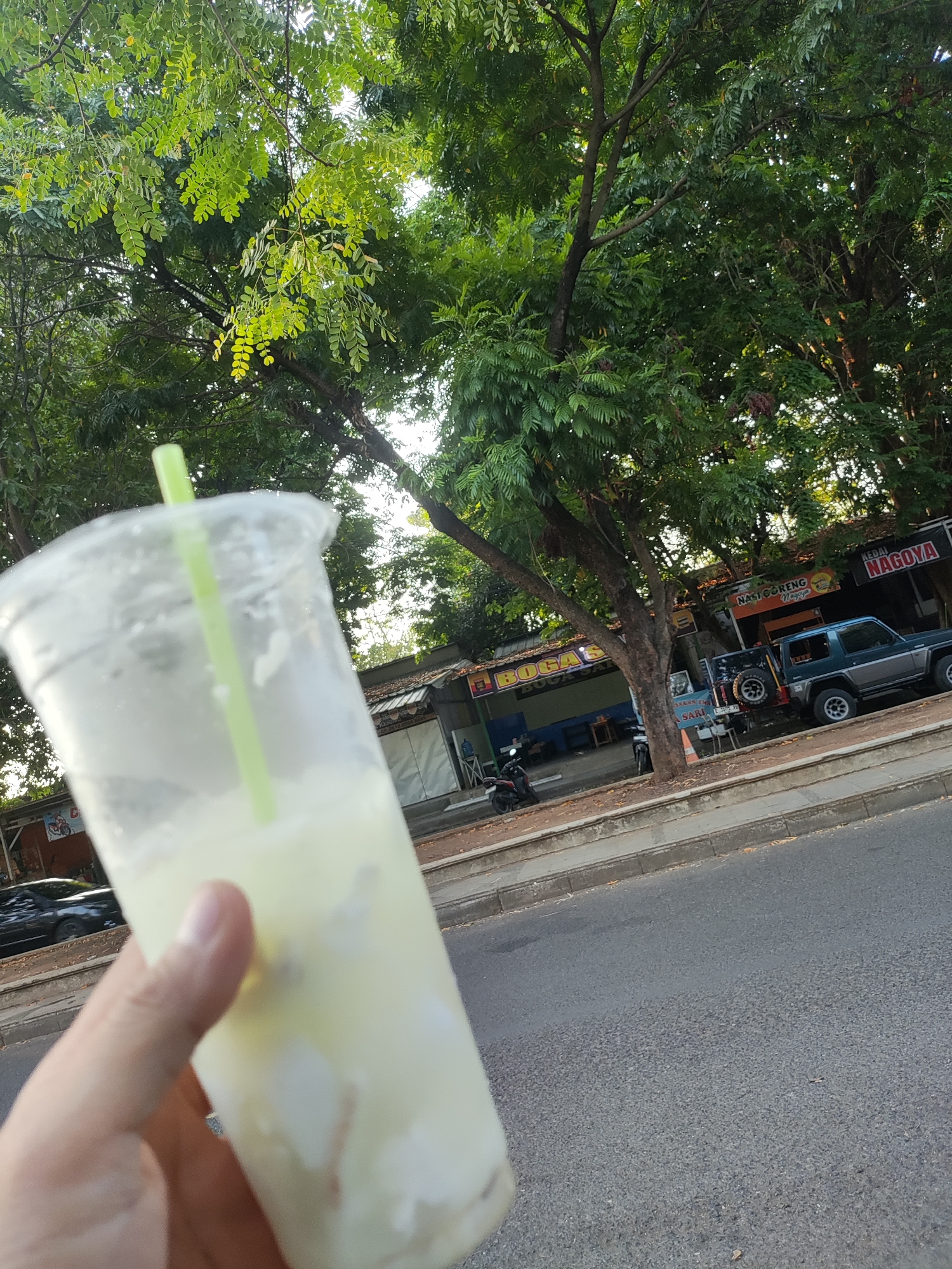 The image shows a clear plastic cup with a light-colored beverage and a green straw, held in a person's hand. In the background, there is a street with a sidewalk and several small shops. A dark blue SUV is parked on the side of the road, and a motorcycle is visible near one of the shops. Trees with lush green foliage line the street, suggesting an outdoor setting. Signs on the shops indicate they are food establishments, with one sign reading "Nasi Goreng Nagoya" and another "Kedai Nagoya". The presence of these shop names and the general urban streetscape suggest the location is likely Cirebon, Indonesia, given that "Nagoya" is a common food stall name in the region. The overall atmosphere appears to be a casual, possibly afternoon or early evening scene.