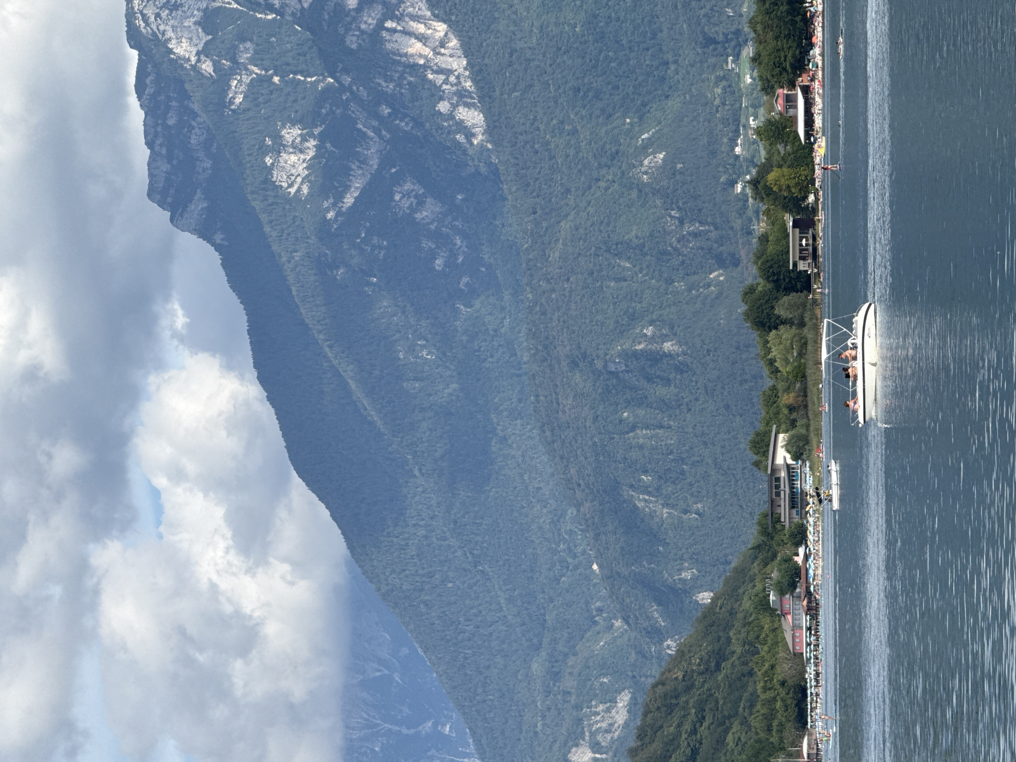 The image captures a scenic view of Calceranica al Lago, Italy, on a cloudy day. A vast, dark blue lake occupies the foreground, with gentle ripples on its surface. Several boats are visible, including a white speedboat with several people on board, a smaller boat, and another boat in the distance. The shoreline in the mid-ground features a lively beach scene with numerous beach umbrellas and sunbathers. Buildings, possibly hotels or restaurants, are situated along the shore, backed by a dense green forest. Towering in the background are majestic, tree-covered mountains, with rocky outcrops and lighter colored rock formations visible on their slopes. The sky above is filled with dramatic, grey and white clouds.