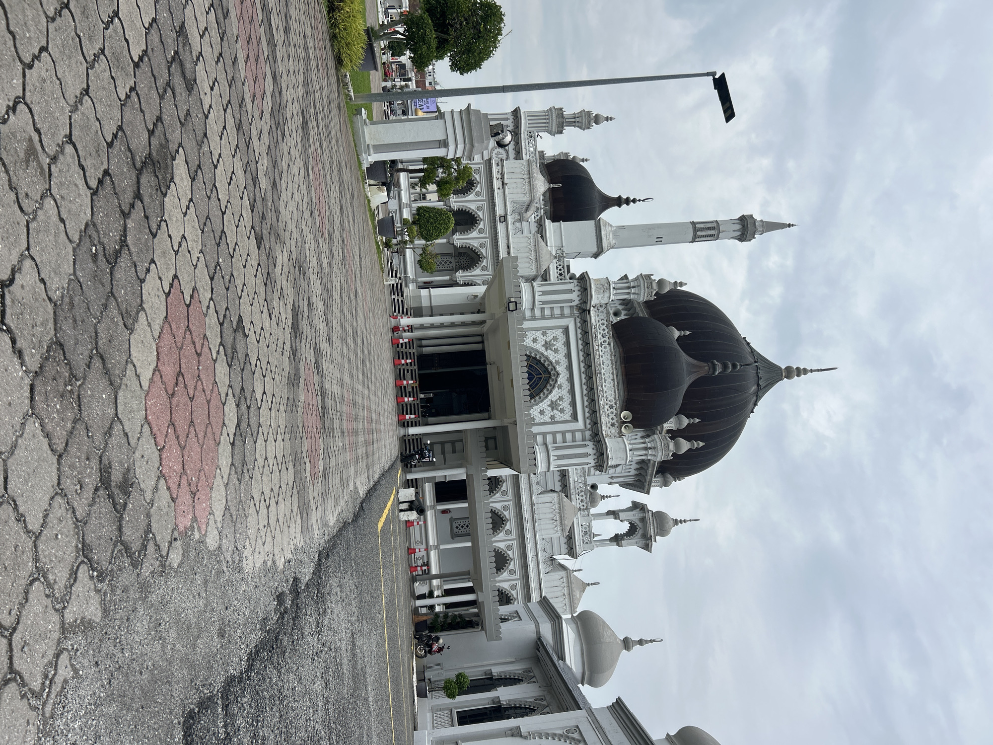 This image shows the exterior of the Zahir Mosque in Alor Setar, Malaysia. The mosque is a prominent landmark, characterized by its white facade, multiple domes, and a tall minaret. The foreground features a patterned paved walkway, with asphalt to the right. Several orange traffic cones are visible along the edge of the walkway, suggesting a temporary barrier or designated area. Two motorcycles are parked near the entrance of the mosque. The sky is overcast, indicating a cloudy day. Trees and some potted plants are also present on the grounds. In the background, the urban environment of Alor Setar can be glimpsed, with buildings and a street visible. The perspective of the image is from a low angle, looking up towards the grand architecture of the mosque.