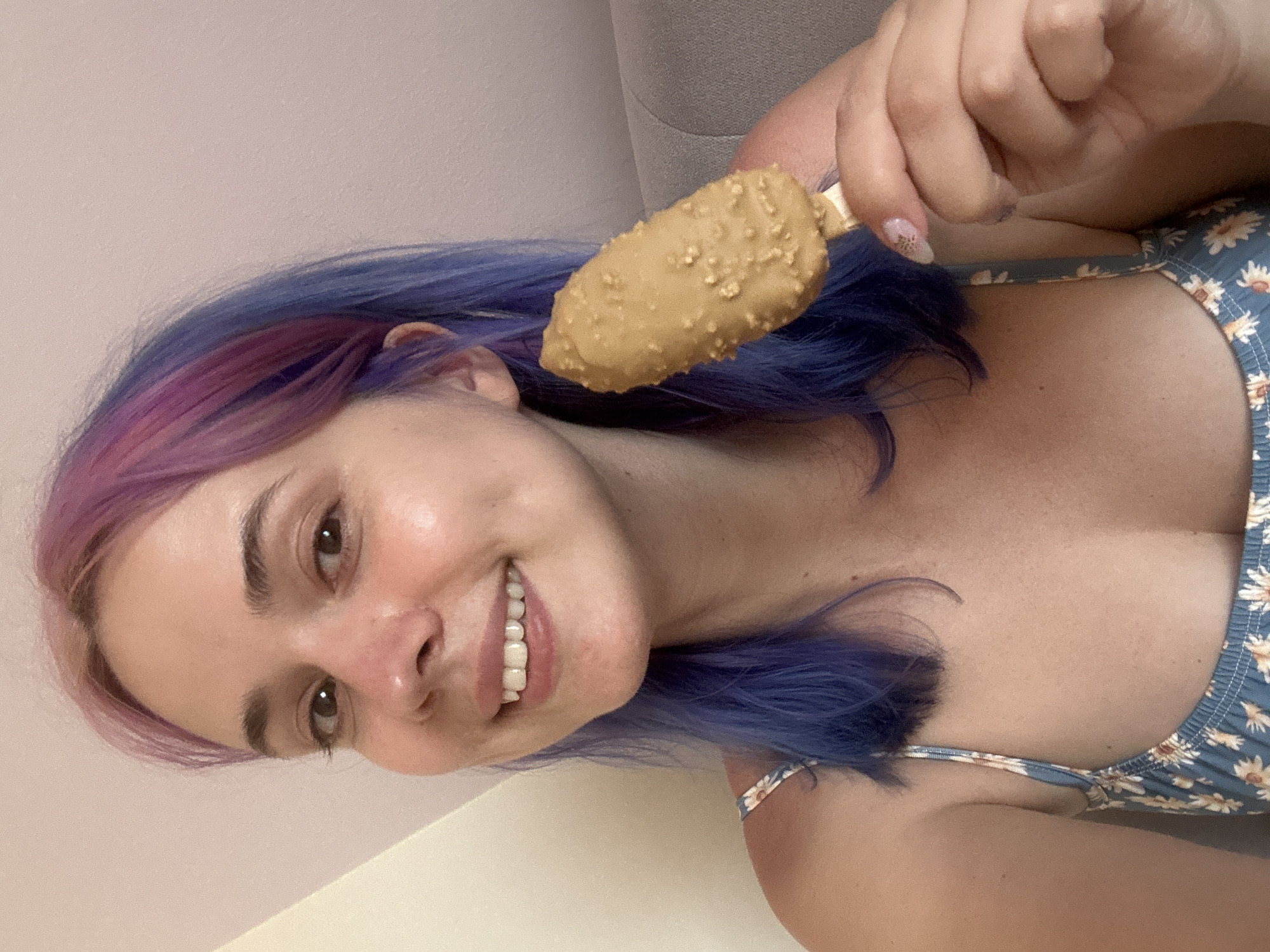 A woman is holding a tan, nugget-covered ice cream bar in her right hand. She has purple and blue hair, fair skin, and is wearing a light blue floral print top. She is smiling and looking towards the camera. The background consists of light beige walls and a gray sofa or cushion. The location appears to be indoors, but there are no specific indicators to pinpoint the location to Gijón/Xixón, Spain.