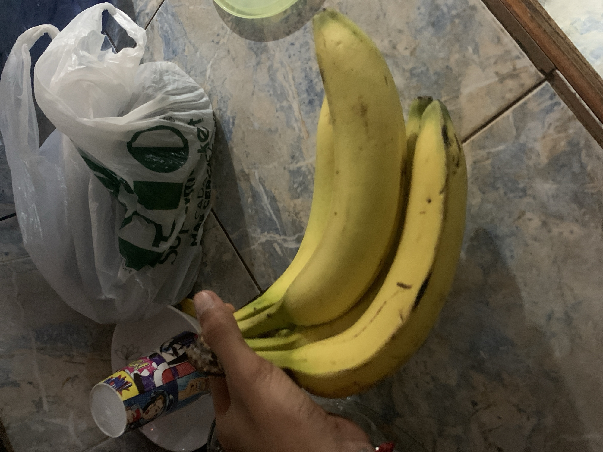 A hand holds a bunch of three ripe bananas above a white, character-printed disposable cup on a small white saucer. Beside them, there is a partially visible white plastic grocery bag with green printing. The items are on a tiled surface with a light blue and grey marbled pattern. The environment appears to be indoors, likely a kitchen or dining area. No specific location in the city of Santa Ana, Venezuela, can be determined from the image.