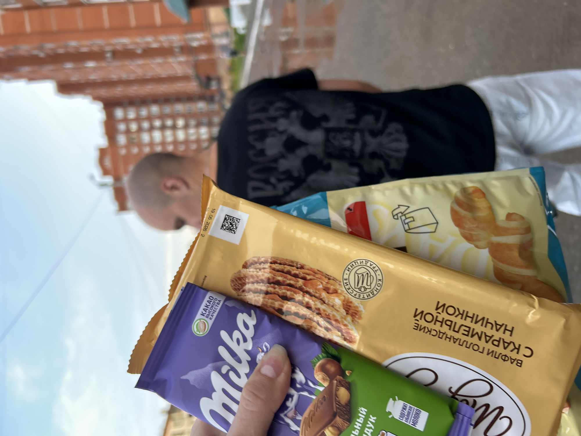 In Chaikovsky, Russia, a person's hand holds up several confectionery items against a blurred backdrop of a residential building and a street with wet pavement. The items include a purple Milka chocolate bar and two packages of wafers. One wafer package is golden yellow with an image of waffles and text in Russian that translates to "Holland Wafers with Caramel Filling." The other visible package appears to be a snack with images of croissants. In the background, a bald man wearing a black t-shirt with a Russian emblem and white shorts walks away from the camera. The sky is visible above.