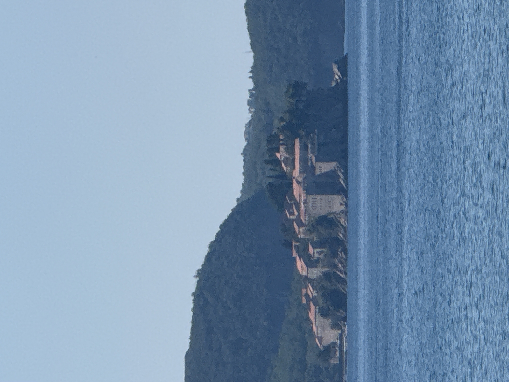 The image shows a view of a coastal village nestled against a steep, tree-covered mountainside, with a body of water in the foreground. The village consists of several buildings with reddish-brown roofs, clustered together along the mountainside. The architecture suggests a Mediterranean or Balkan setting. The water appears to be calm with a gentle ripple effect on the surface. The sky is a clear, pale blue, indicating a bright, sunny day, likely in the late morning or early afternoon. The overall scene evokes a sense of tranquility and natural beauty. The location is identified as Rafailovići, Montenegro. There is no visible text in the image.