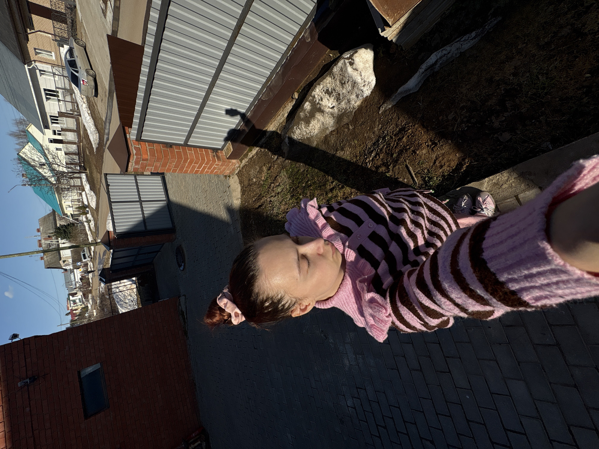 A medium-angle, outdoor shot shows a woman in a pink and brown striped cardigan in the Russian city of Прикамский. The subject is light-skinned with brown hair. She is on a gray brick patio with sunlight shining on her. The patio borders a yard with dirt, grass, and a rock. A gray metal garage-like door is to the left of the yard. Houses and a car are across the street. A brown brick building is to the left of the patio.