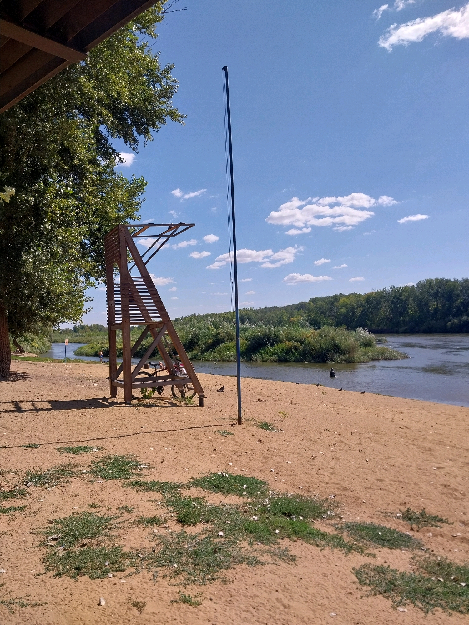 The image depicts a sandy beach next to a calm river on a sunny day with scattered white clouds. On the beach, there is a wooden lifeguard tower with a bicycle leaning against its base. A person is visible in the river, standing in the water. The riverbank is lined with dense green trees and vegetation. In the foreground, the sand is sparsely covered with green grass. The scene appears to be a recreational area, possibly within the city of Orenburg, Russia, given the natural landscape.