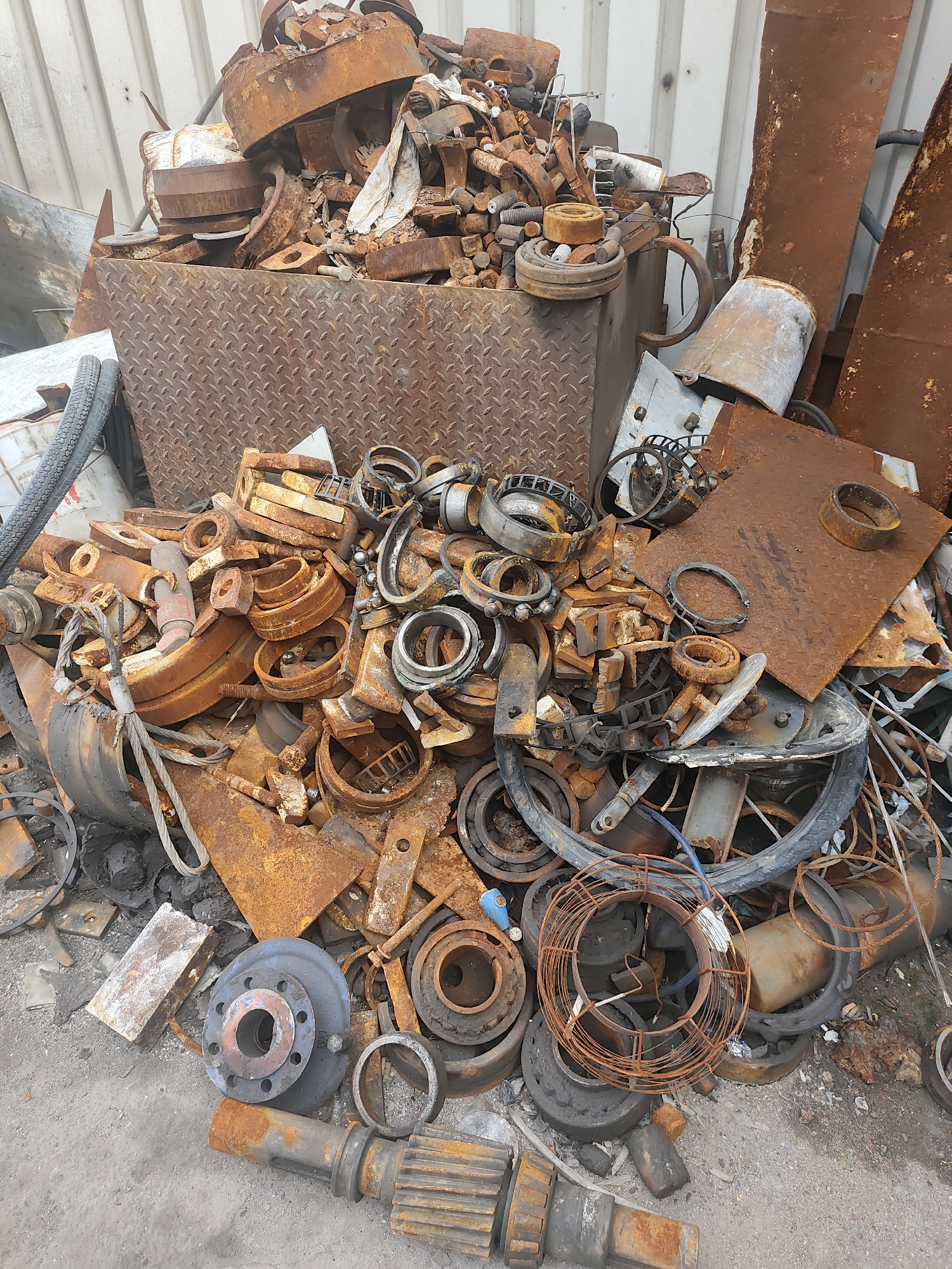 This image shows a pile of rusted metal scrap, including various gears, bearings, pipes, and other machine parts, located in an outdoor industrial or junkyard setting. The scrap is densely packed and covers the ground, with some pieces forming a significant mound. The background consists of corrugated metal siding, typical of a warehouse or industrial building. The ground appears to be a mixture of dirt and gravel. There are no people or animals visible in the image. The scene suggests a location for the collection or processing of scrap metal. This could be a scrap yard in the city of Police, Poland, given the context.